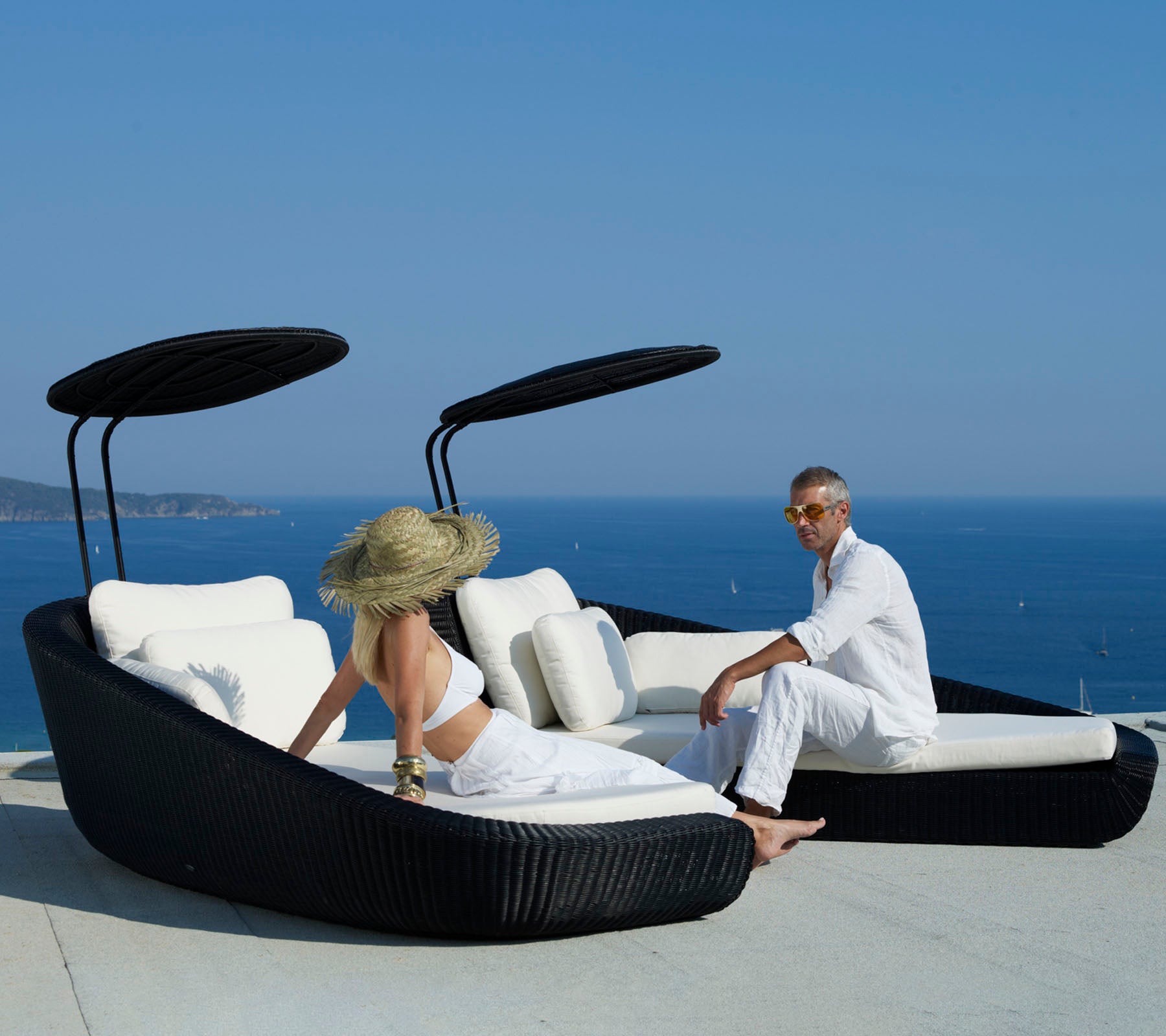 Deux personnes se détendent sur un lit de soleil courbé et élégant avec une vue imprenable sur l'océan, offrant un design noir épuré et de douces coussins blancs.
