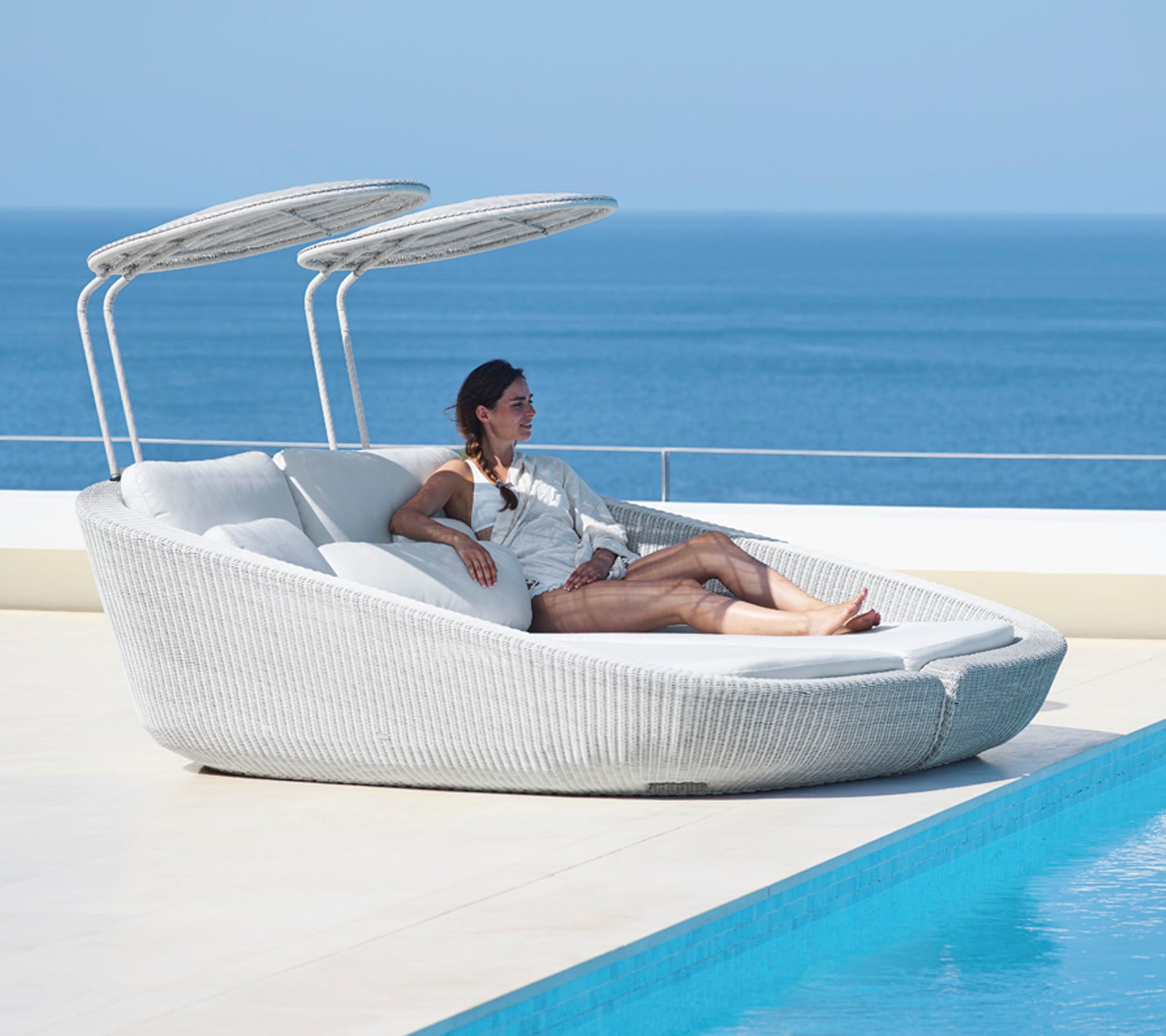 Une femme se détend sur un fauteuil lounge moderne et élégant avec des auvents réglables à côté d'un plan d'eau tranquille.