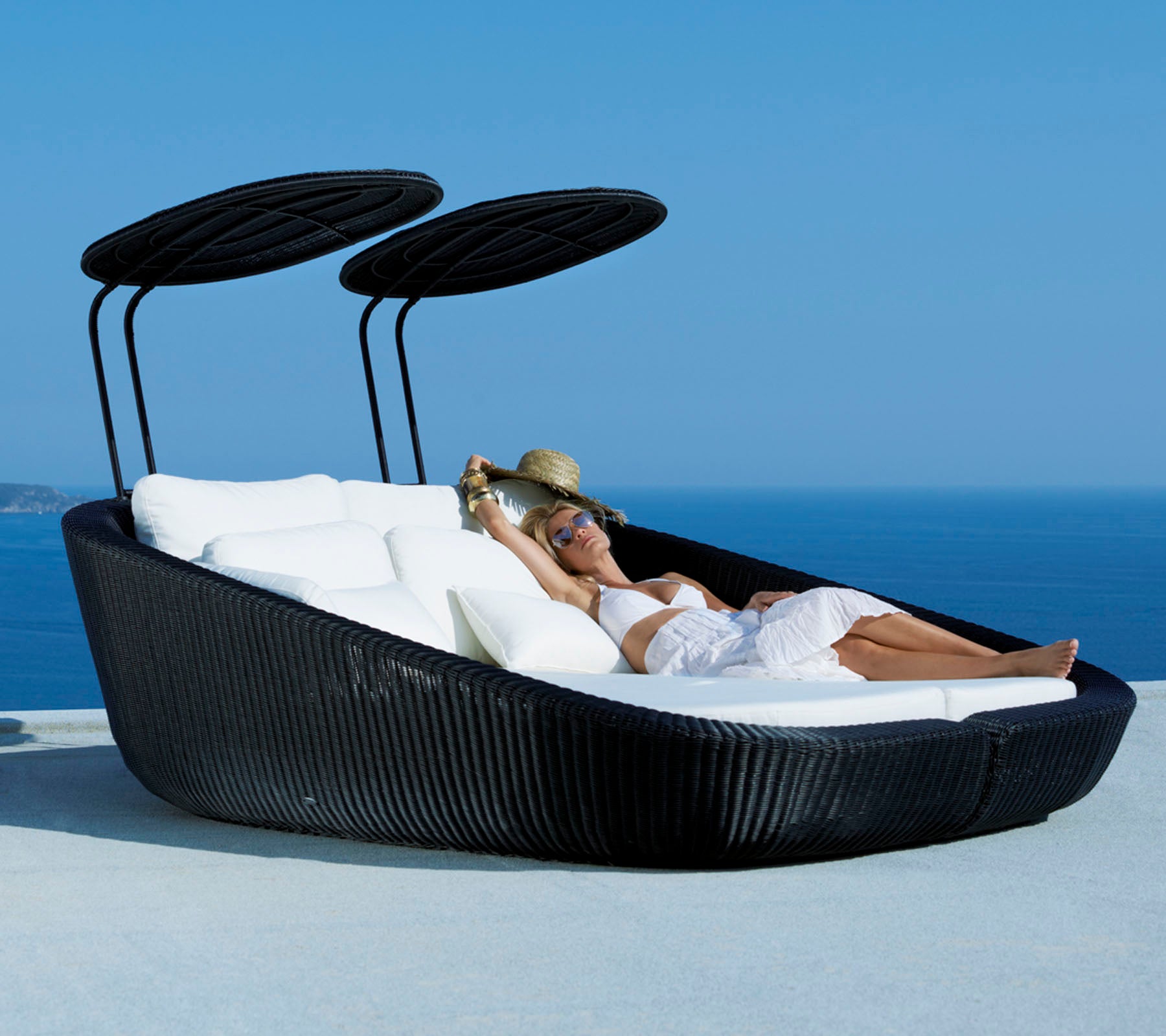 Un lit de lounge extérieur élégant avec un design épuré, doté de coussins doux et de auvents ajustables, sur fond de paysage océanique pittoresque.