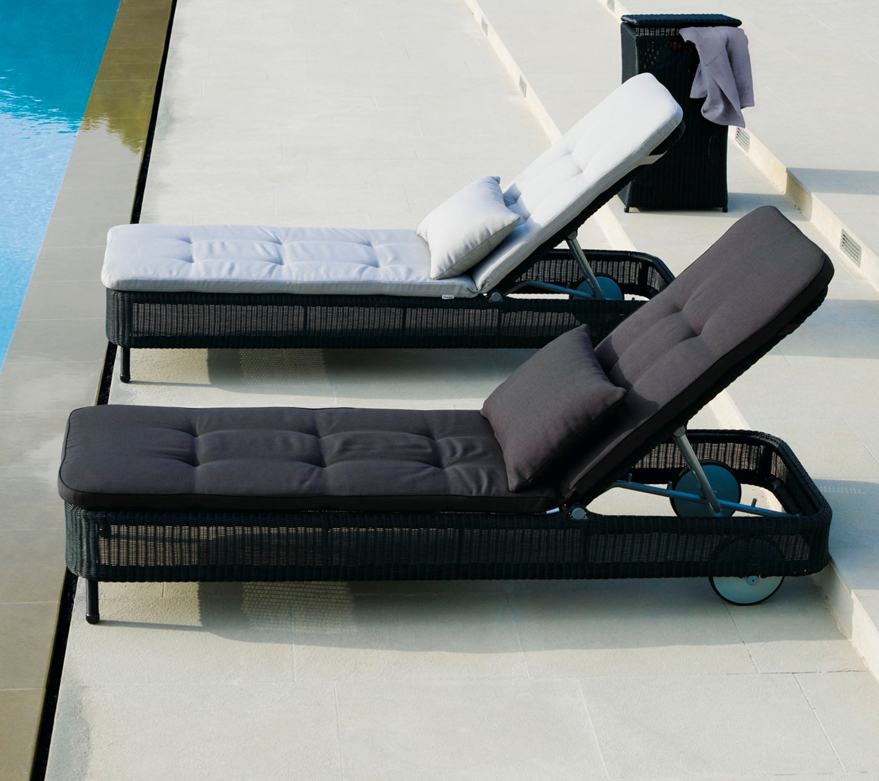 Deux chaises longues de couleurs contrastées, l'une en gris clair avec un coussin, et l'autre en gris foncé, positionnées à côté d'une piscine.