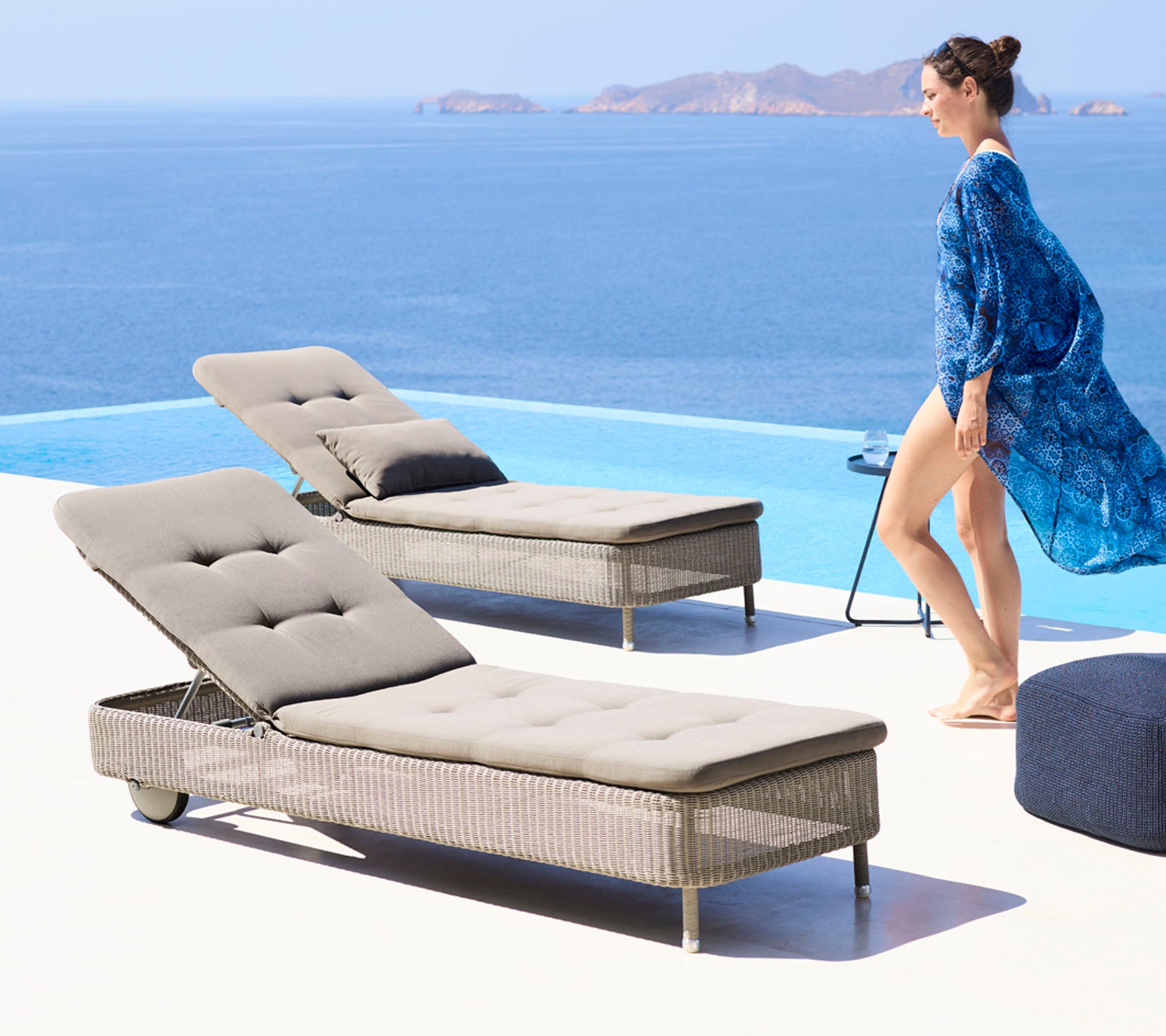 Une femme dans une couverture bleue fluide marche à côté de deux chaises longues élégantes près d'une piscine, surplombant une vue océane sereine.