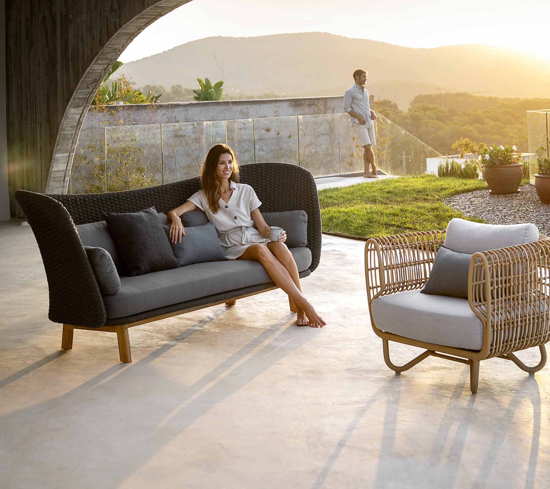 Un aménagement de sièges élégant avec une femme se relaxant sur un canapé moderne avec divers coussins, et une chaise à proximité, situé devant un arrière-plan pittoresque.