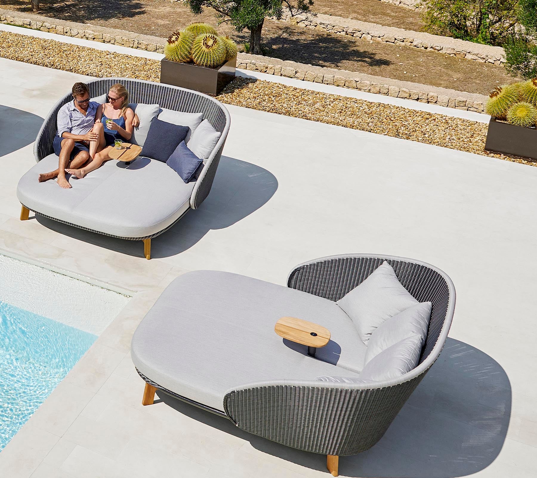 Deux personnes se détendent sur des chaises longues extérieures élégantes près d'une piscine, profitant de leur temps libre avec des coussins et une petite table.