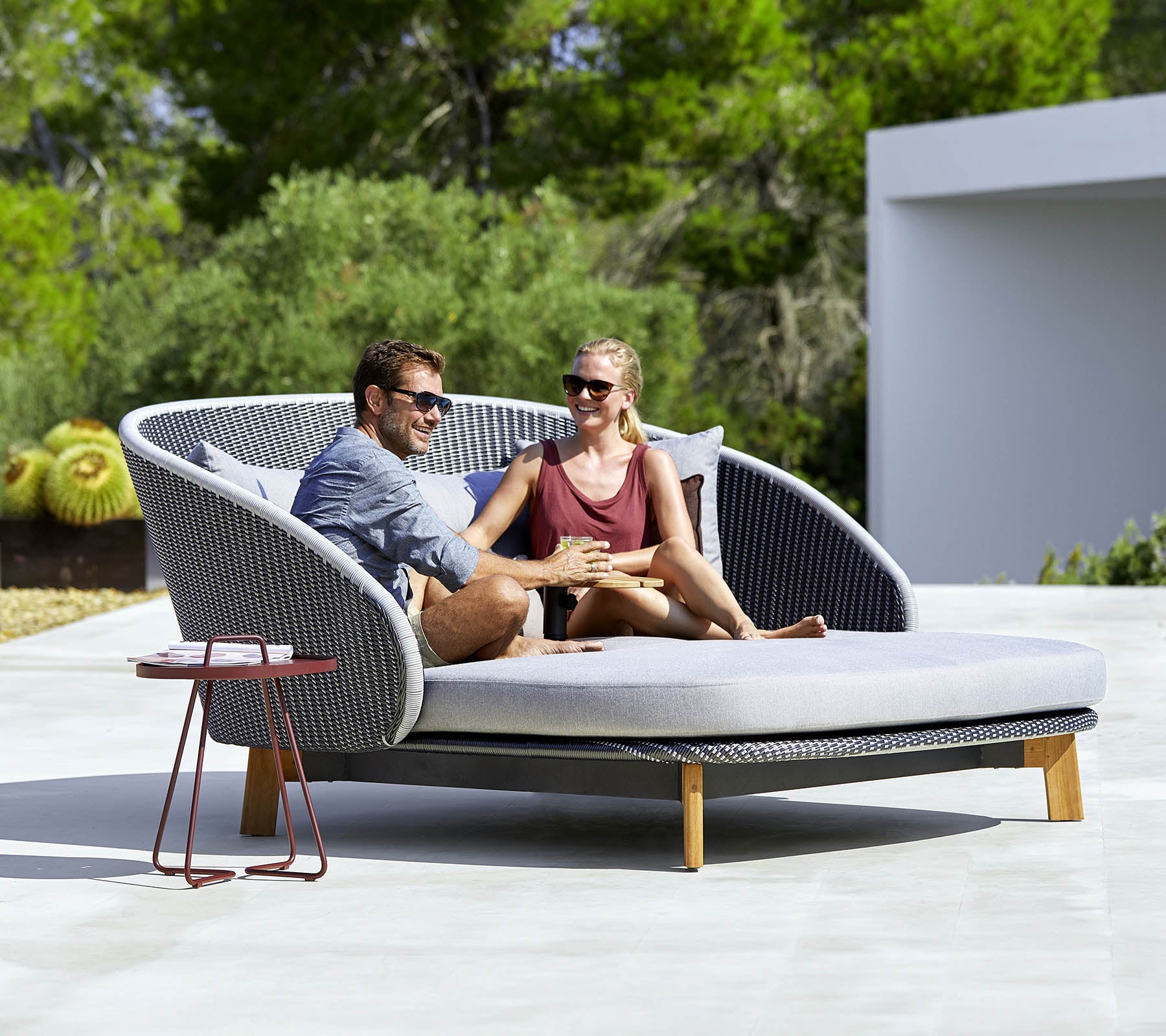 Un canapé d'extérieur élégant et courbé avec deux personnes se relaxant ensemble, entouré de verdure et d'une petite table d'appoint à proximité.