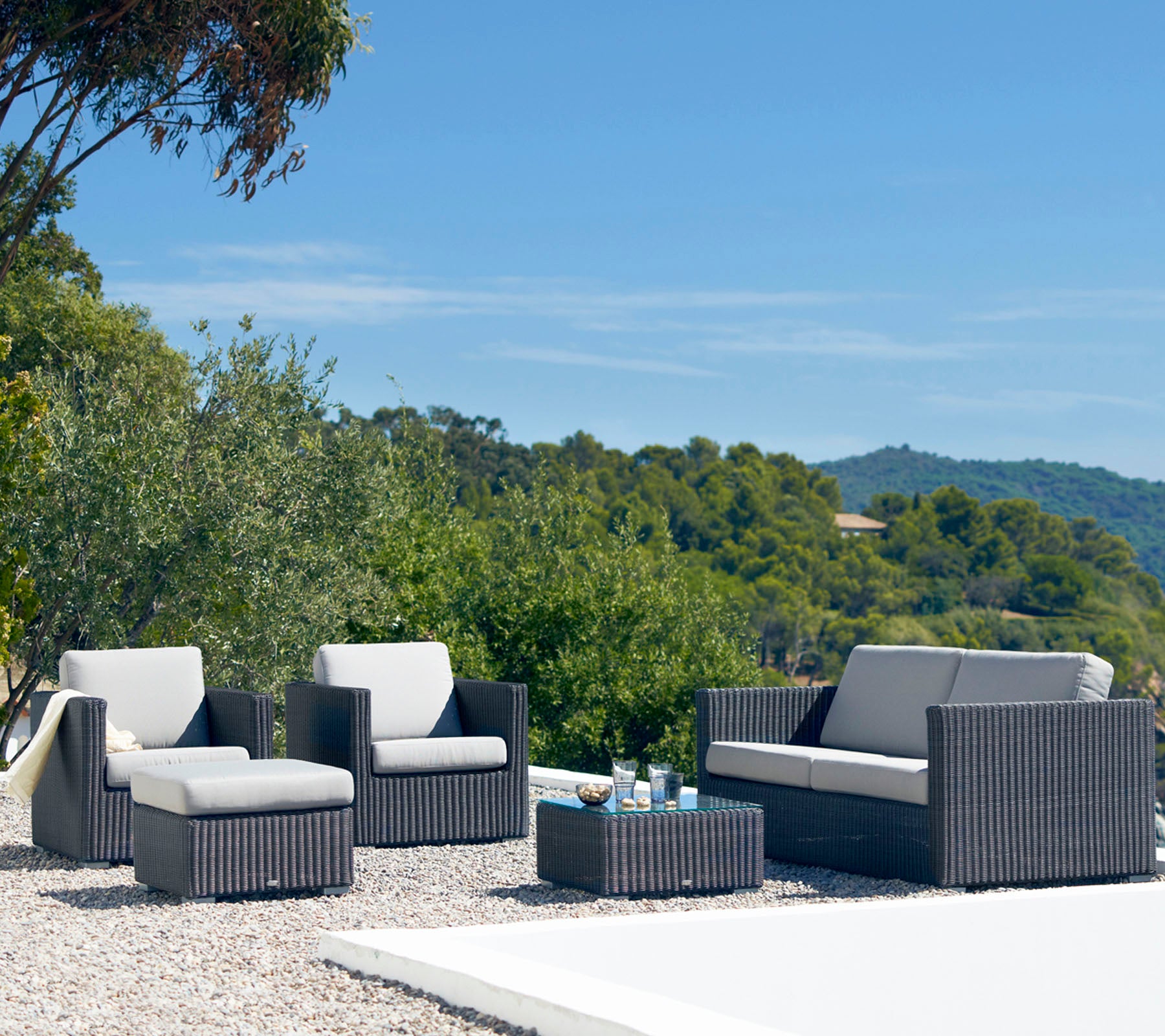 Ensemble de mobilier d'extérieur élégant comprenant un canapé, deux fauteuils et une table basse au design épuré et avec des coussins neutres, entouré de verdure.
