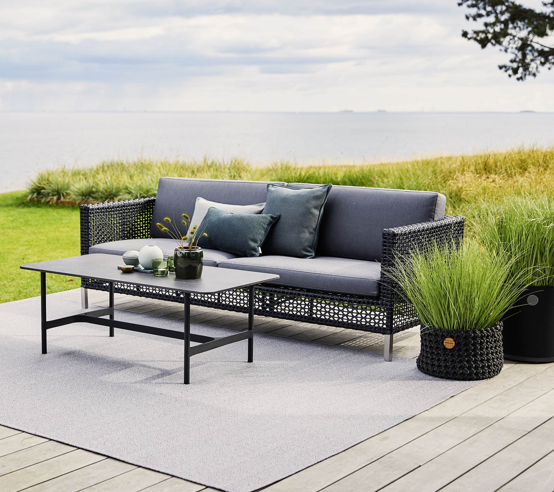 Des sièges d'extérieur élégants avec des coussins gris, une table en verre et des plantes en pot, installés devant un paysage pittoresque.