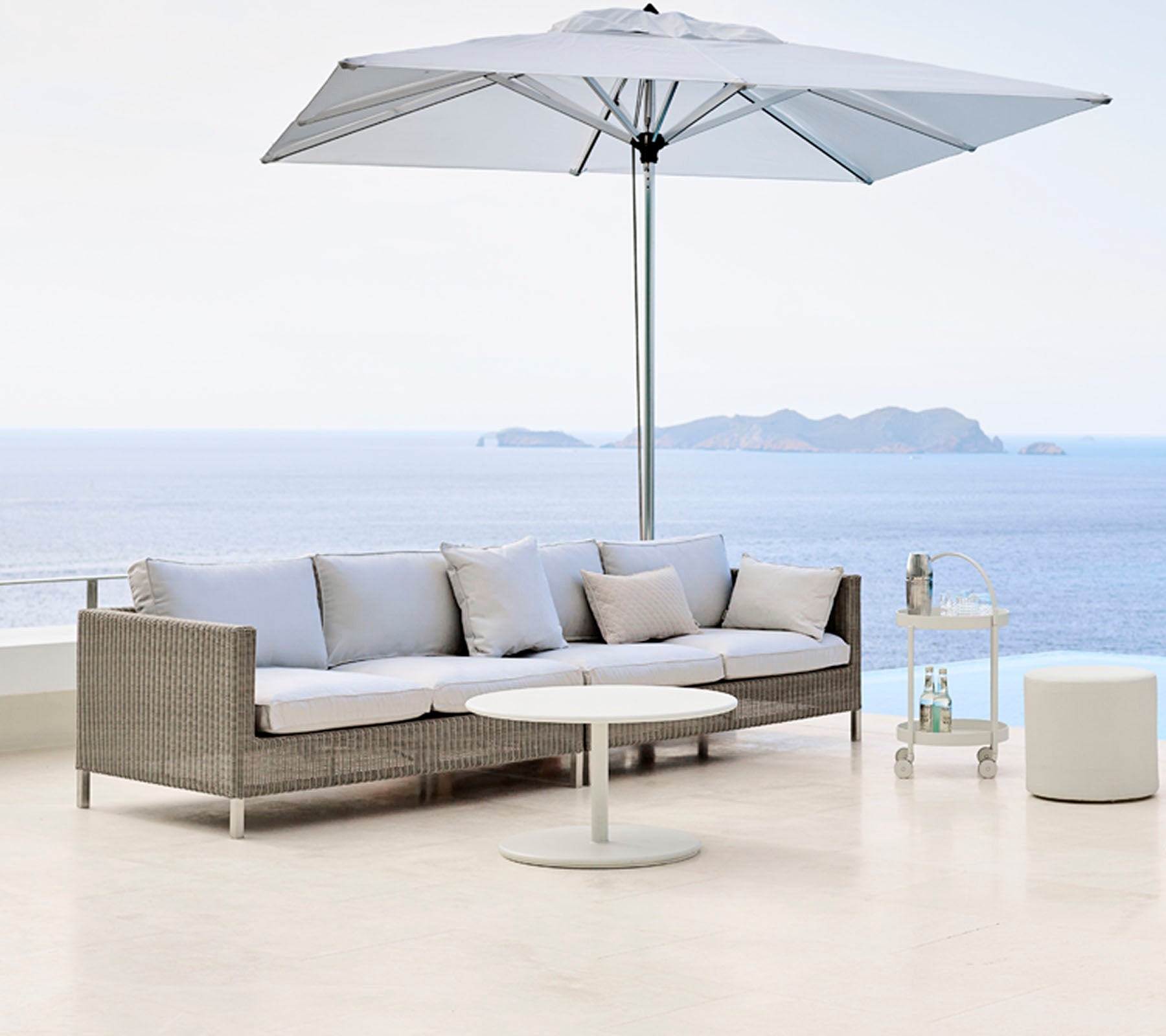 Espace extérieur élégant avec un canapé gris clair, une table ronde et un grand parapluie, surplombant un plan d'eau serein.
