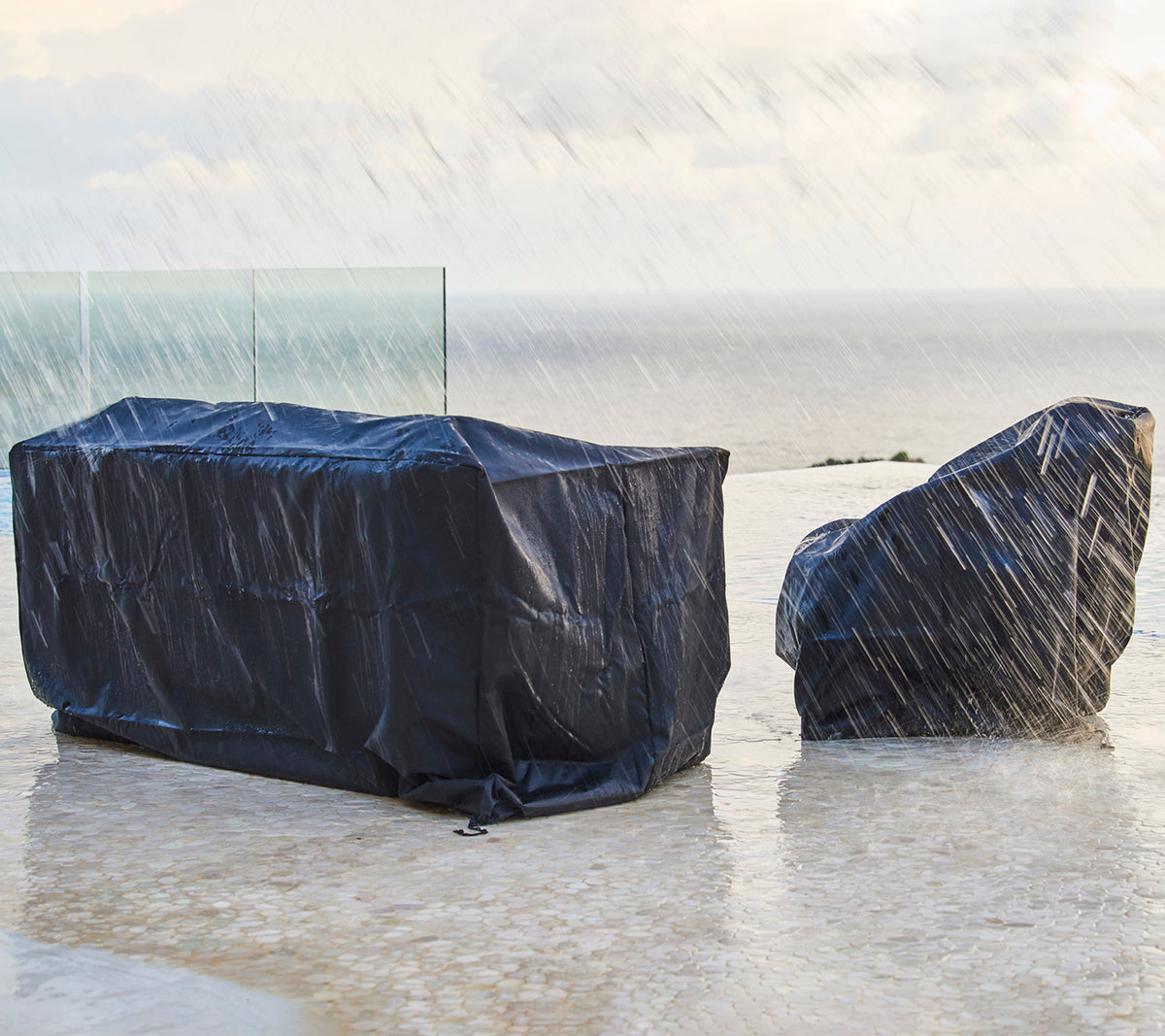 Mobilier d'extérieur couvert de housses de protection sombres, à l'abri de la pluie, avec un arrière-plan d'un ciel nuageux et de l'océan.