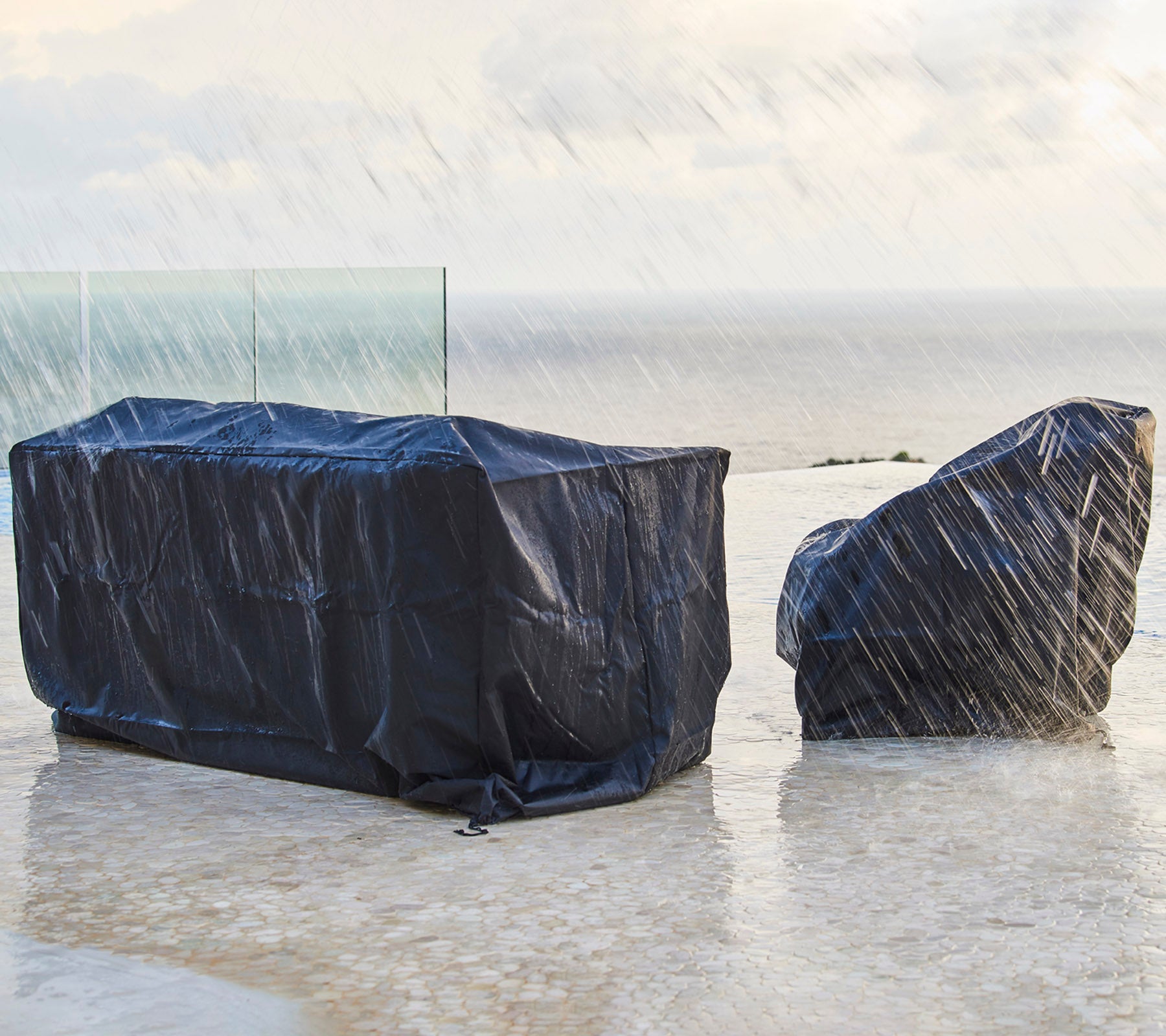 Mobilier d'extérieur couvert de housses de protection sombres, à l'abri de la pluie, avec un arrière-plan d'un ciel nuageux et de l'océan.