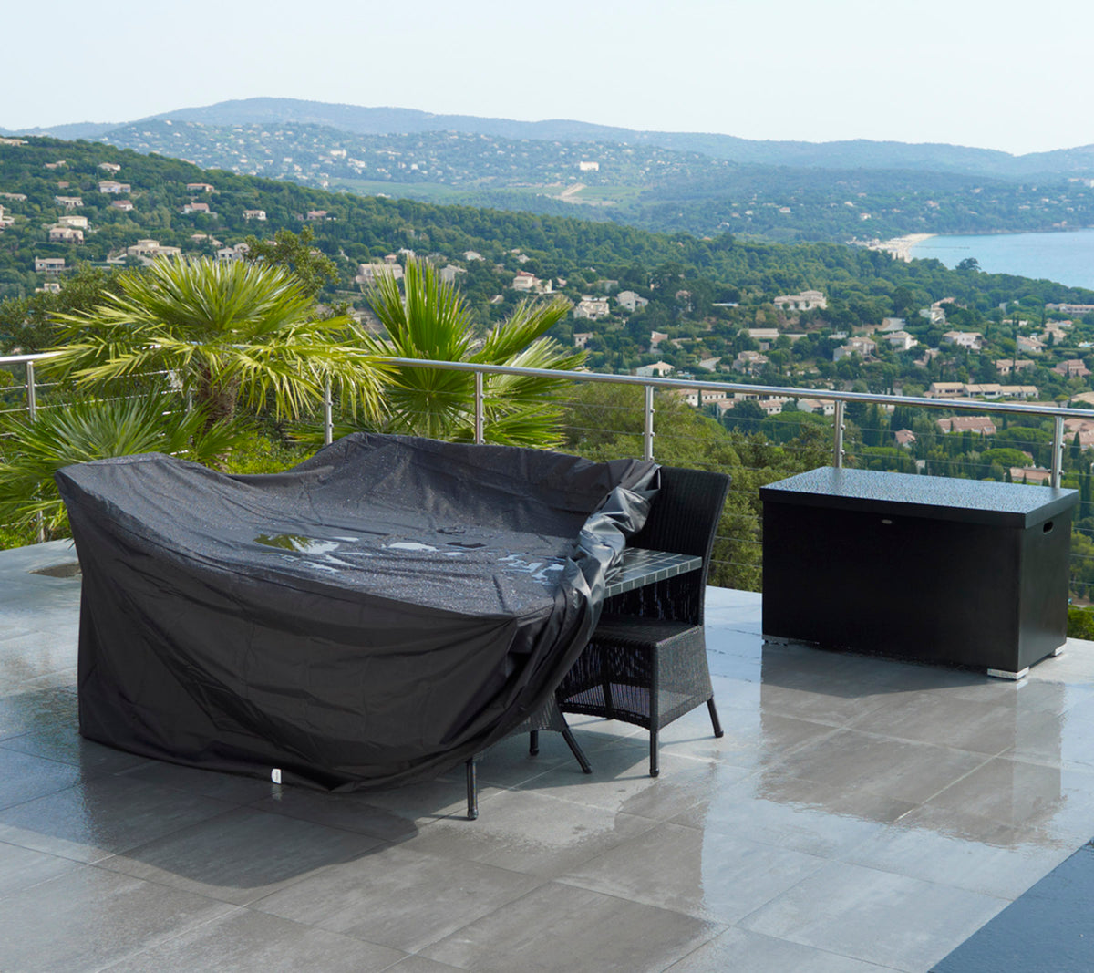 Zone de sièges extérieurs couverte d'une housse de protection noire, accompagnée d'une boîte de rangement assortie et offrant une vue pittoresque sur les collines et l'eau.
