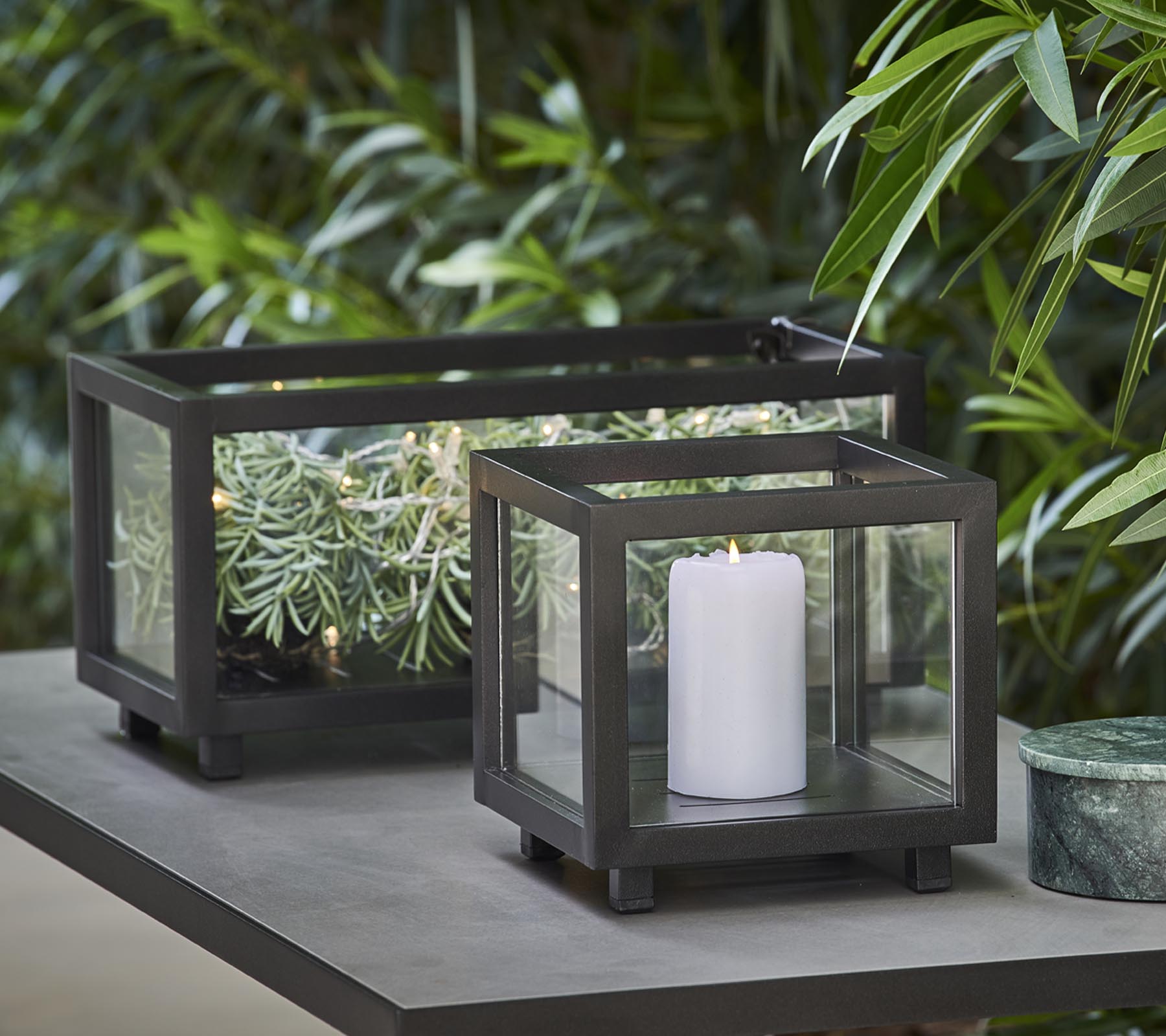 Deux lanternes élégantes avec des panneaux en verre, l'une contenant une bougie blanche et l'autre présentant de la verdure, placées sur une table au milieu du feuillage luxuriant.
