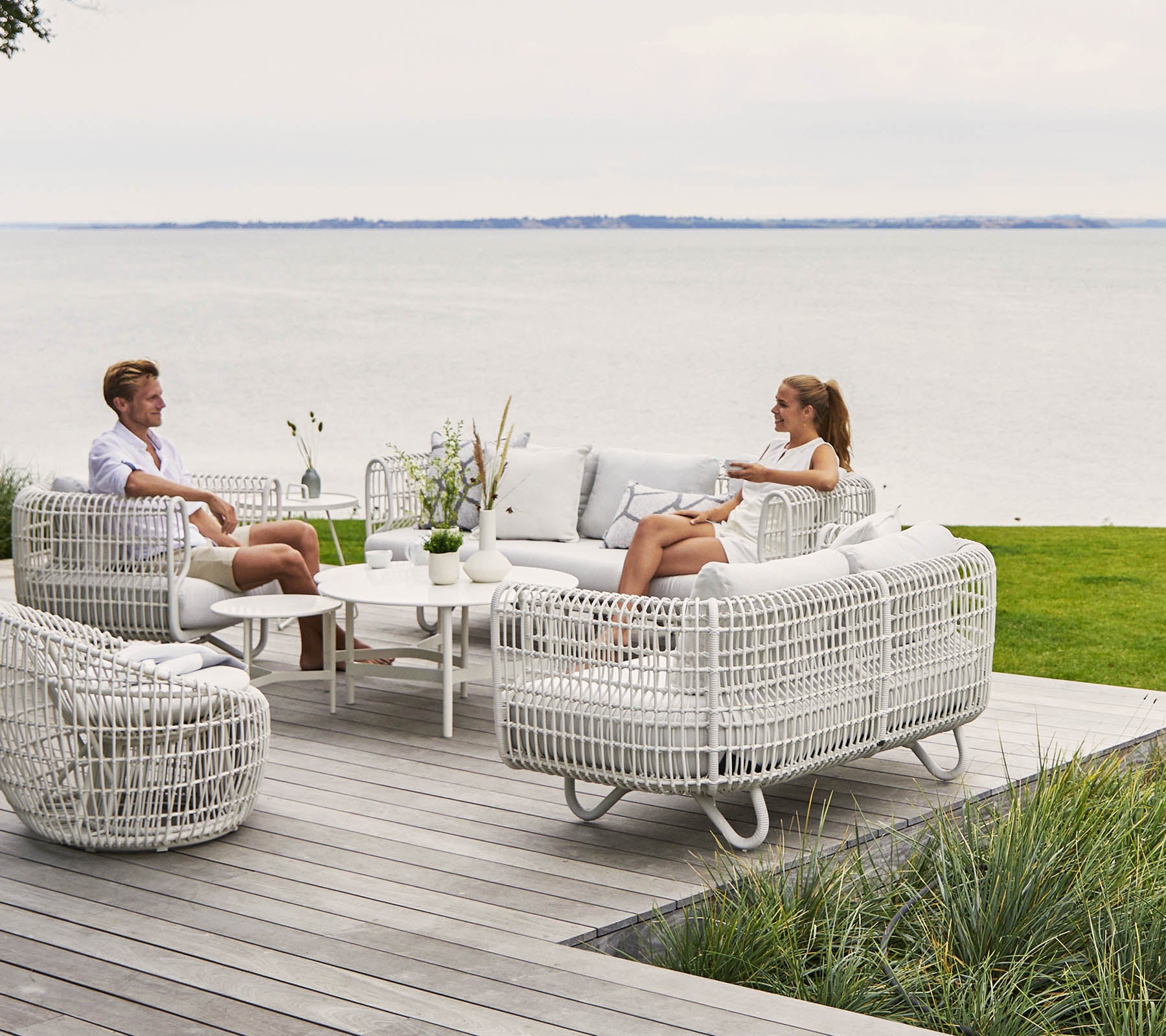 Deux personnes se détendent dans un espace de vie élégant et confortable avec des meubles en osier blanc et une table basse, surplombant une vue sur l'eau pittoresque.