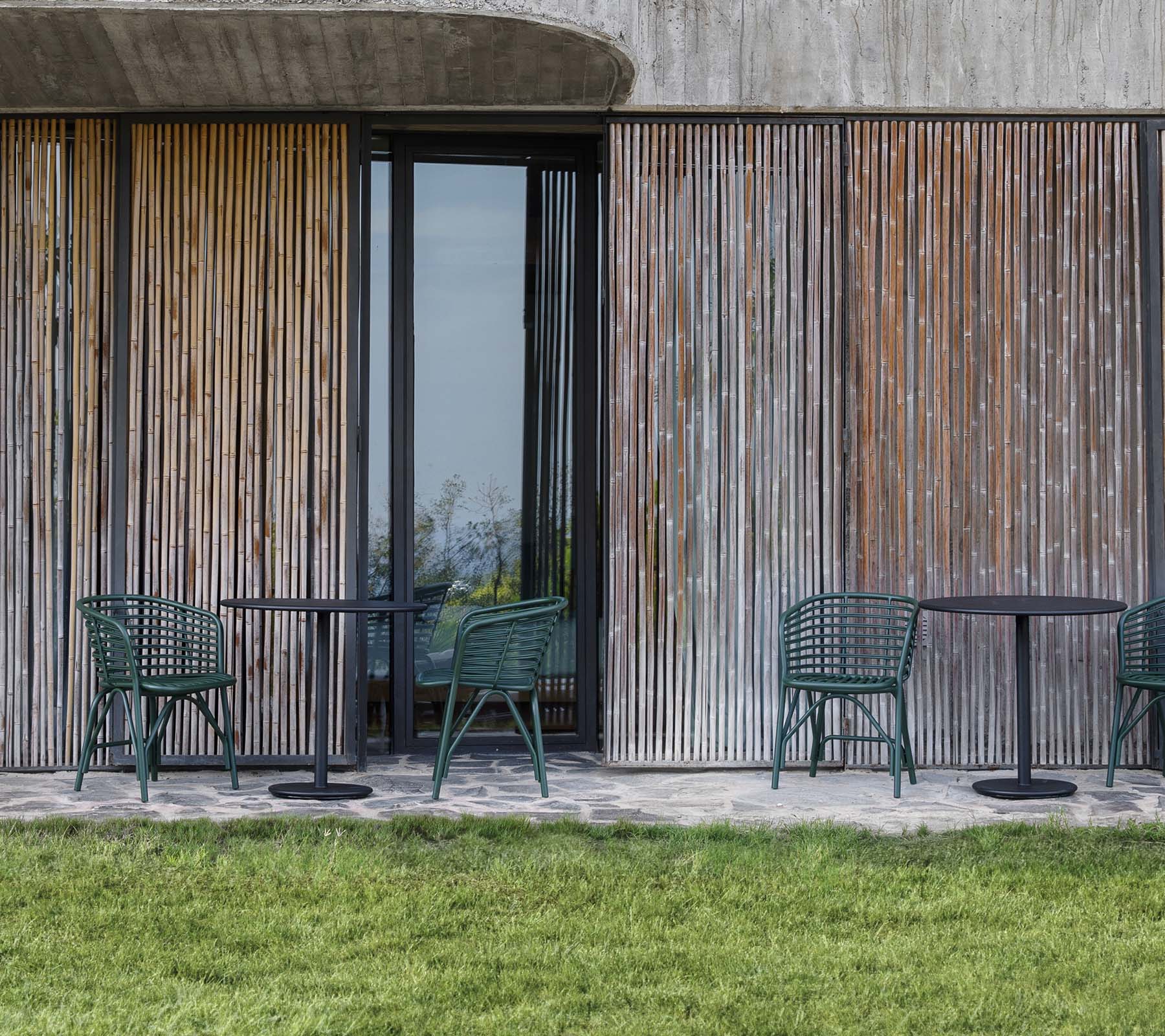 Chaises et tables disposées devant un fond en panneaux de bois, complété par une zone herbeuse.