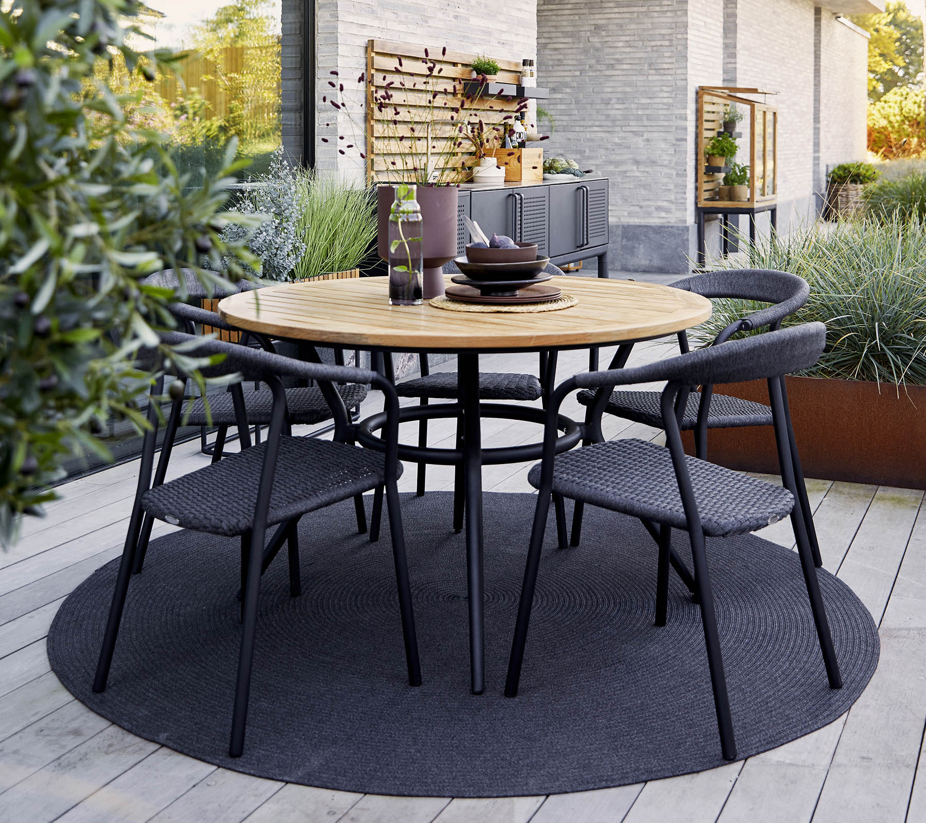 Table ronde en bois avec des chaises noires disposées autour, dressée pour le repas, entourée de verdure et d'un tapis texturé.