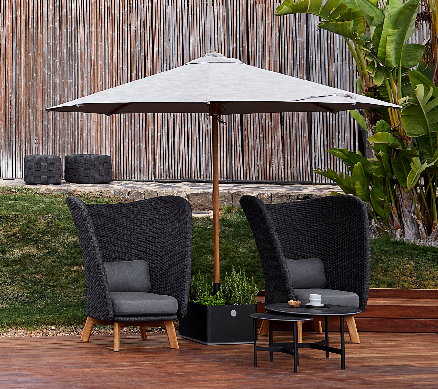Deux chaises noires élégantes avec des coussins gris et une petite table sous un grand parasol, entourées de verdure et d'un sol en bois.