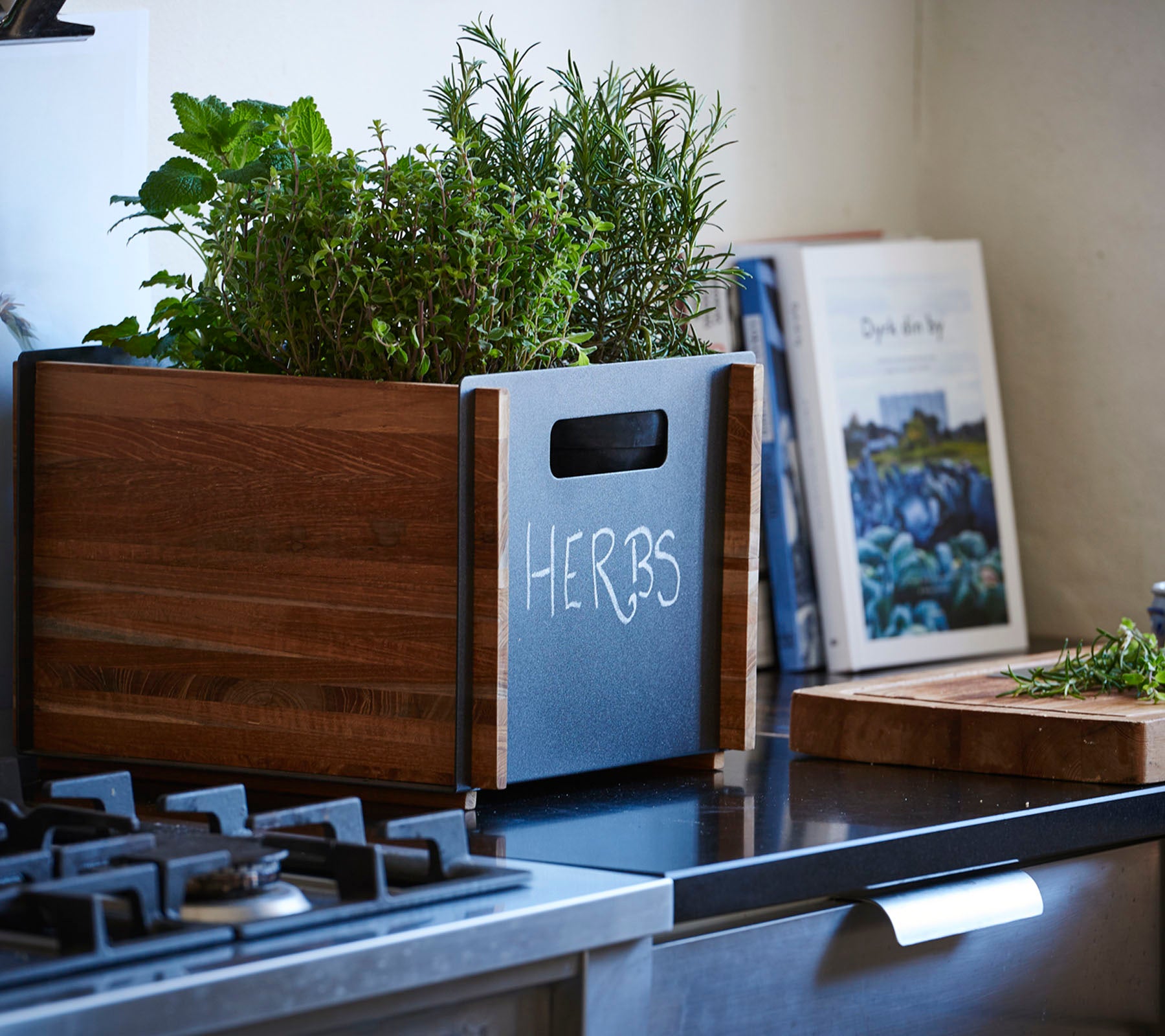 Bac à plantes en bois étiqueté "HERBES," rempli d'herbes fraîches, à côté d'un livre de recettes sur un plan de travail de cuisine.