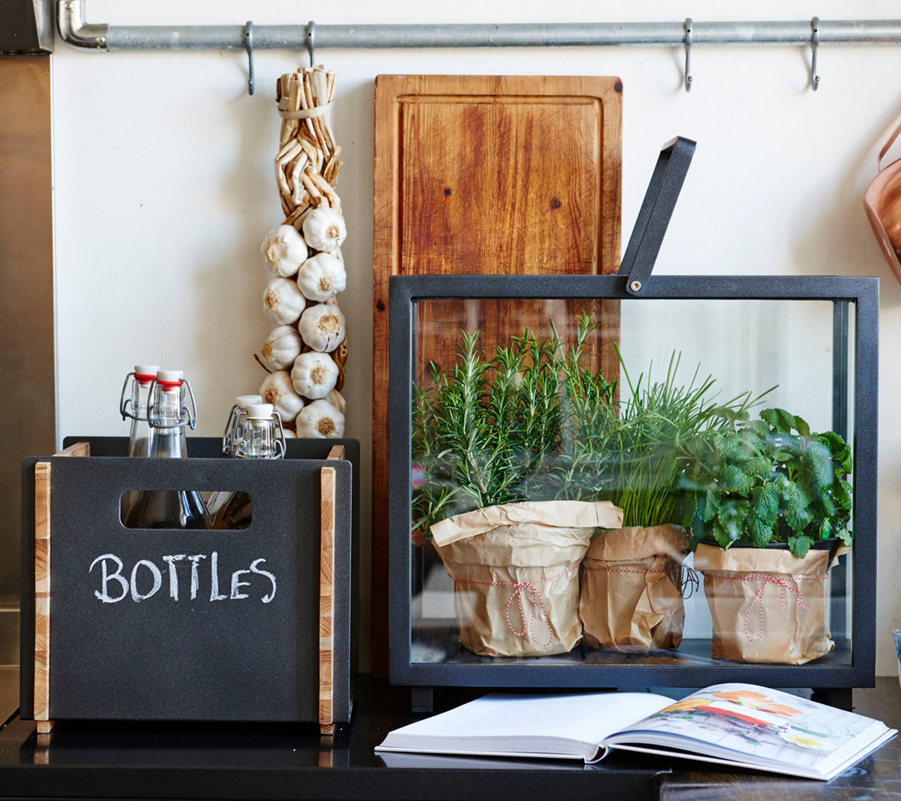 Une vitrine élégante présentant une boîte noire étiquetée 'BOUTEILLES', un conteneur en verre avec des herbes en pot et un livre de cuisine sur une surface.
