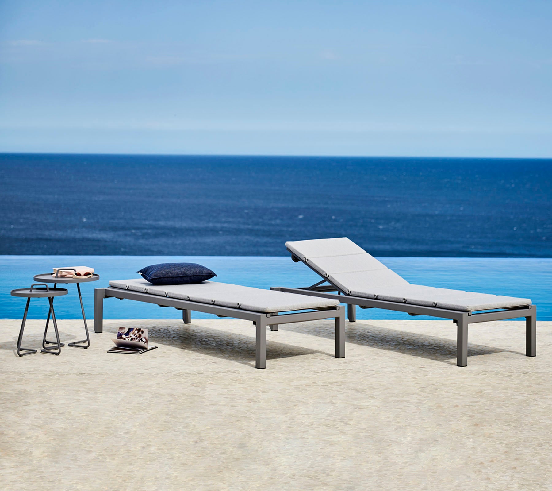 Deux fauteuils confortables au style élégant avec des coussins gris sont placés à côté d'une petite table. En arrière-plan, on aperçoit une vue sereine sur l'océan.