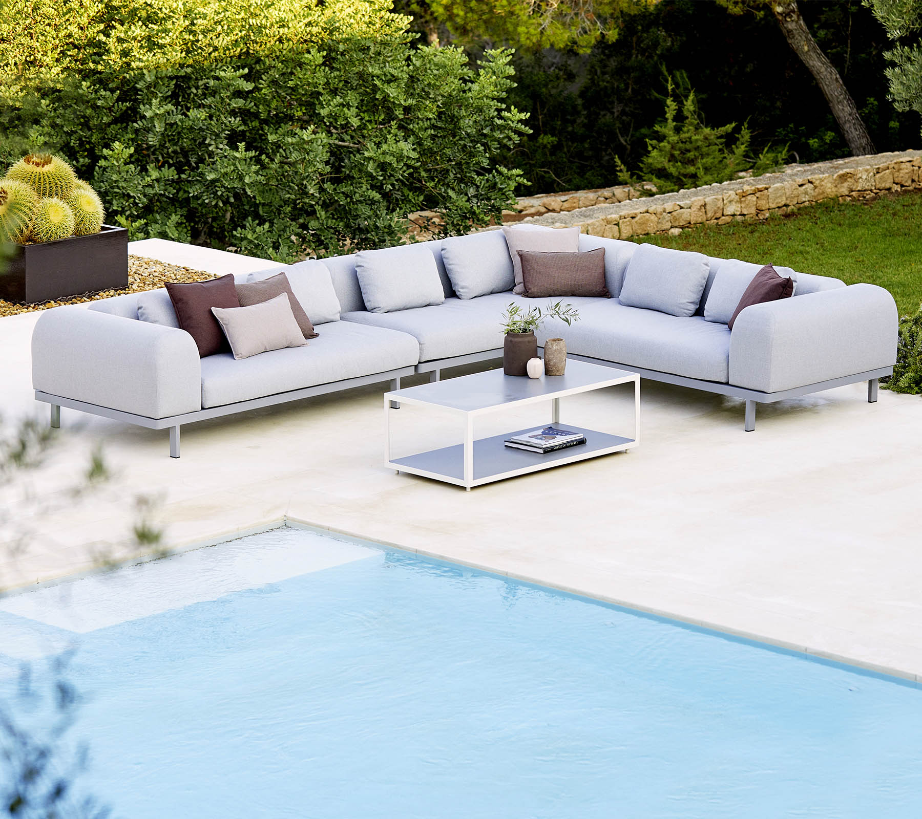 Canapé sectionnel gris clair avec des coussins décoratifs et une table basse en verre, positionné à côté d'une piscine et d'une végétation luxuriante.