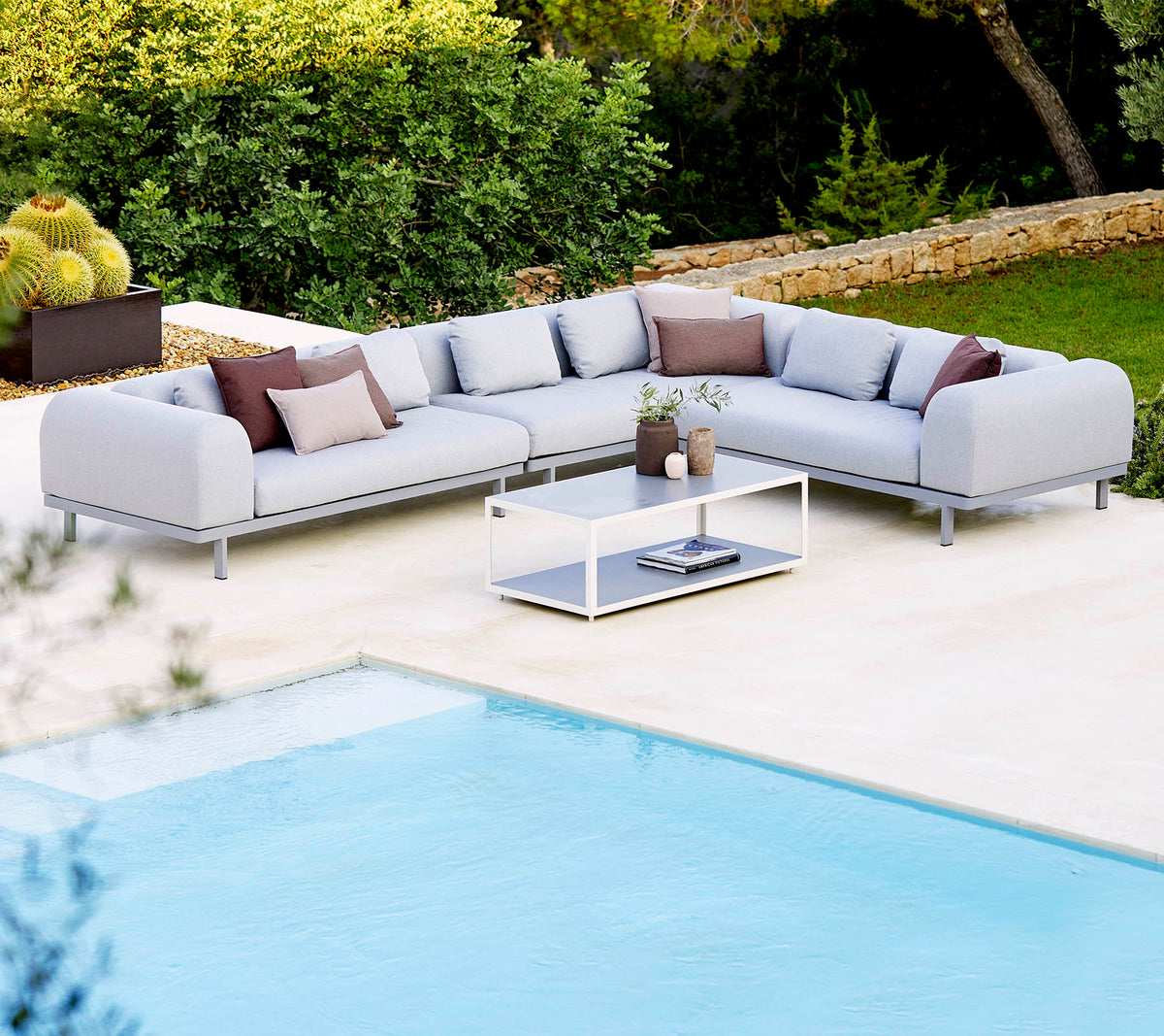 Canapé sectionnel gris clair avec des coussins décoratifs et une table basse en verre, positionné à côté d'une piscine et d'une végétation luxuriante.