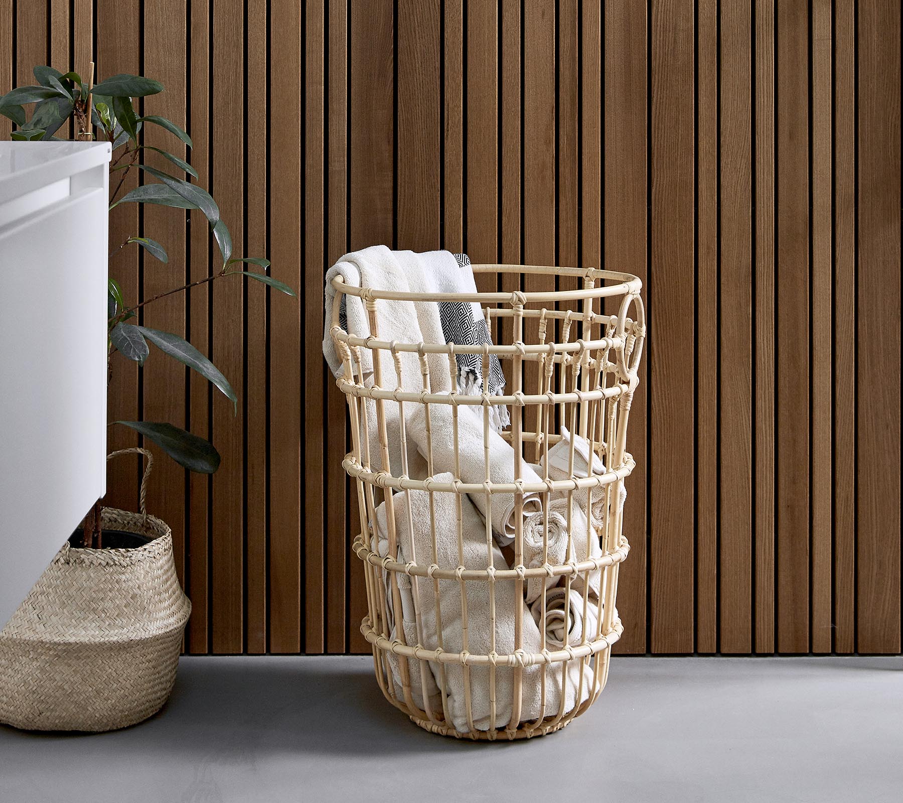 Panier à linge tressé rempli de serviettes roulées, placé à côté d'une plante et d'un mur en bois texturé.