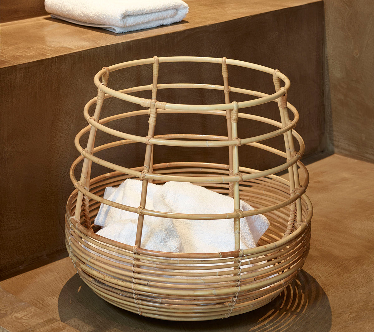 Panier en bambou tressé contenant des serviettes blanches soigneusement empilées, placées sur une surface lisse.