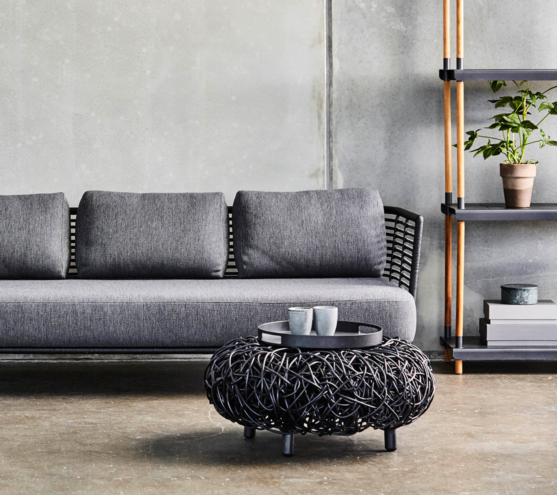 Un canapé gris moderne avec des coussins, associé à une table basse ronde en osier noir et une étagère minimaliste affichant des plantes et de la décoration.