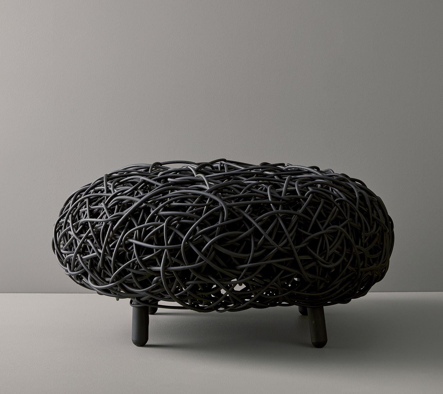 Ottoman tissé noir avec une forme organique et arrondie et un design complexe, reposant sur trois fines jambes contre un fond neutre.