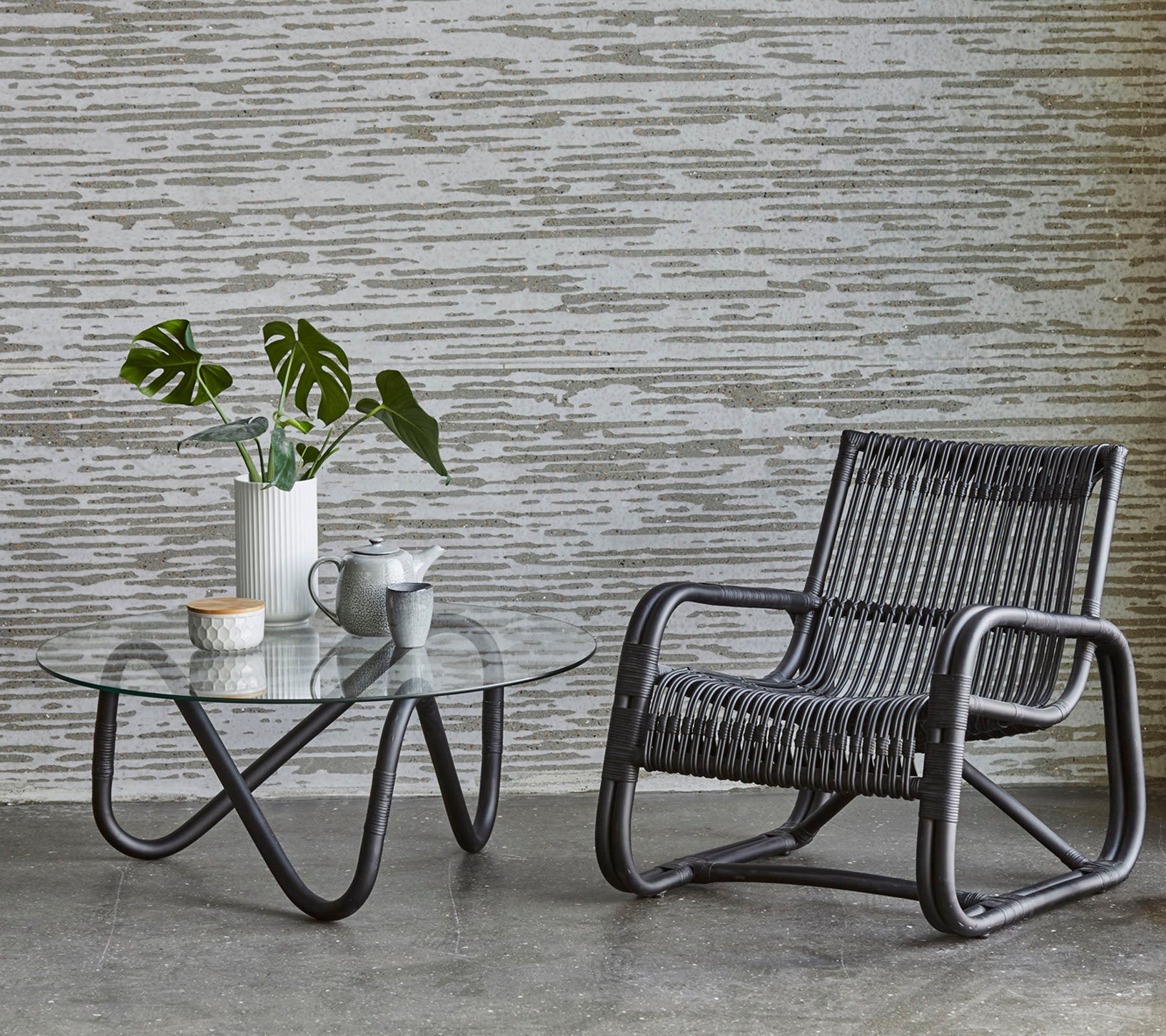 Une chaise élégante à côté d'une table basse en verre, avec un vase de verdure et une petite tasse de thé, contre un mur texturé.