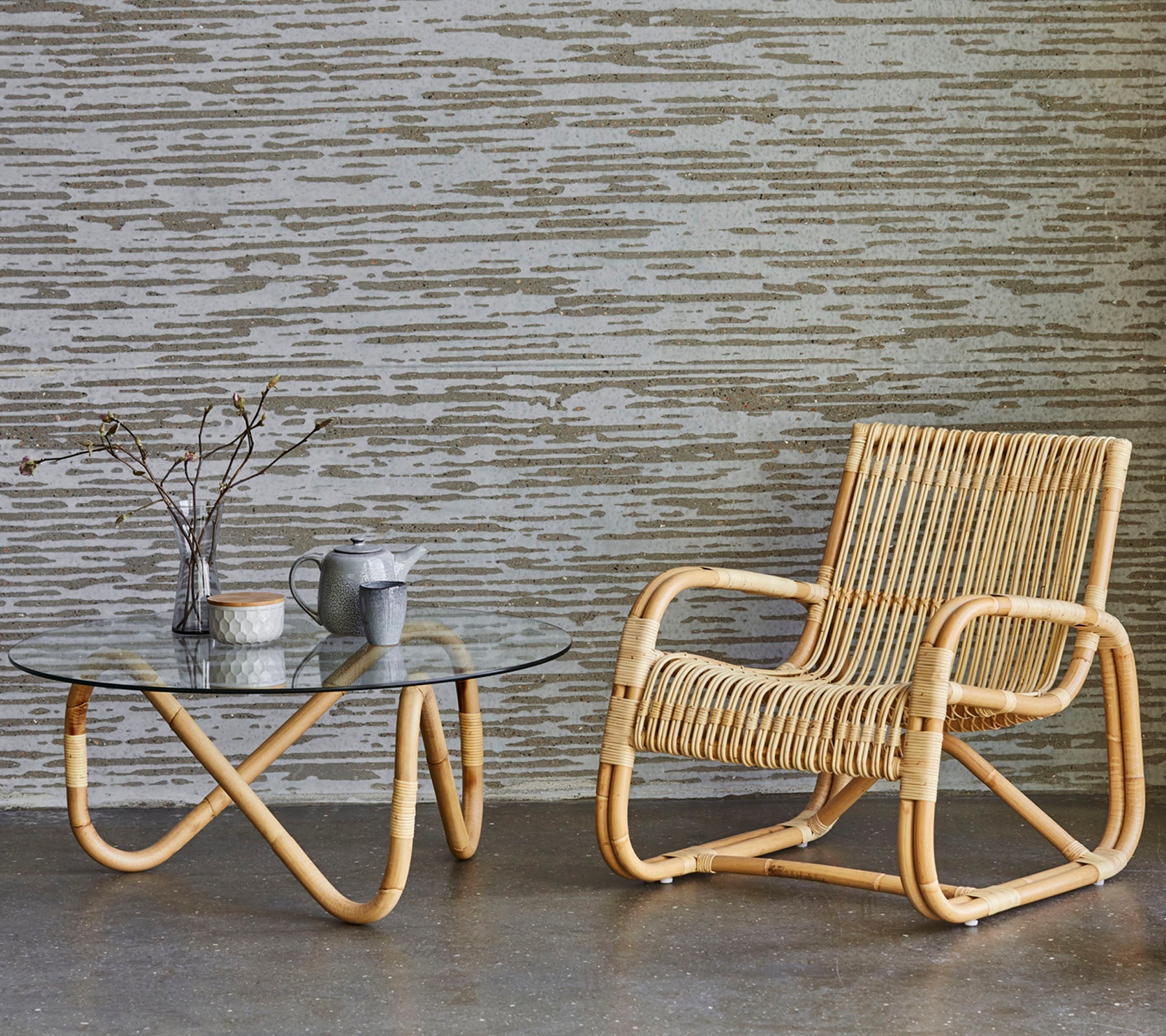 Chaise en rotin et table en verre avec des objets décoratifs, placés contre un mur texturé. Un style de décor minimaliste et moderne.