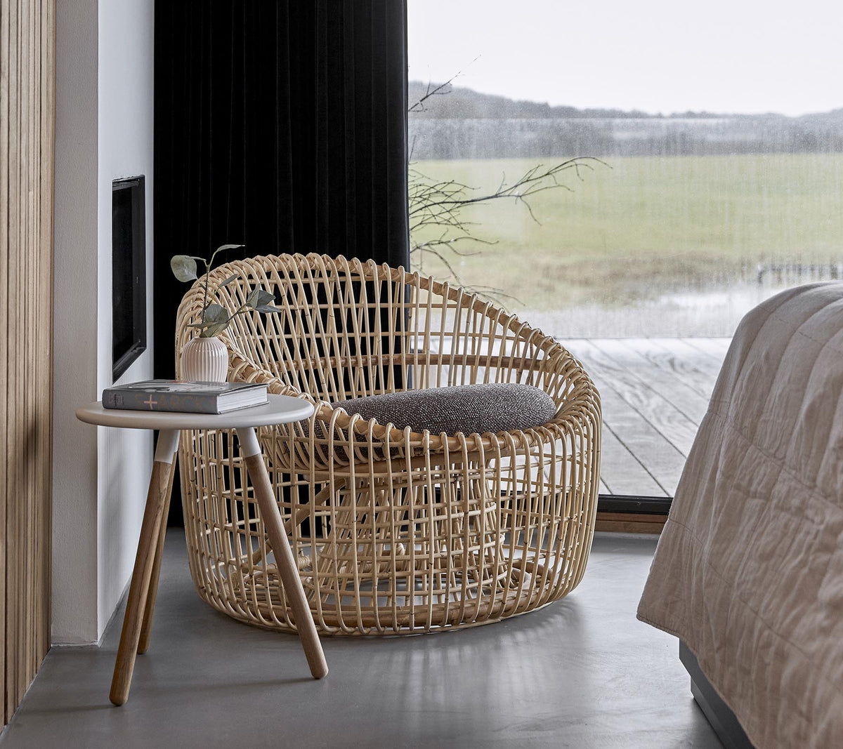 Une chaise tissée élégante accompagnée d'une petite table d'appoint, installée dans un environnement serein avec de grandes fenêtres laissant entrer la lumière naturelle.