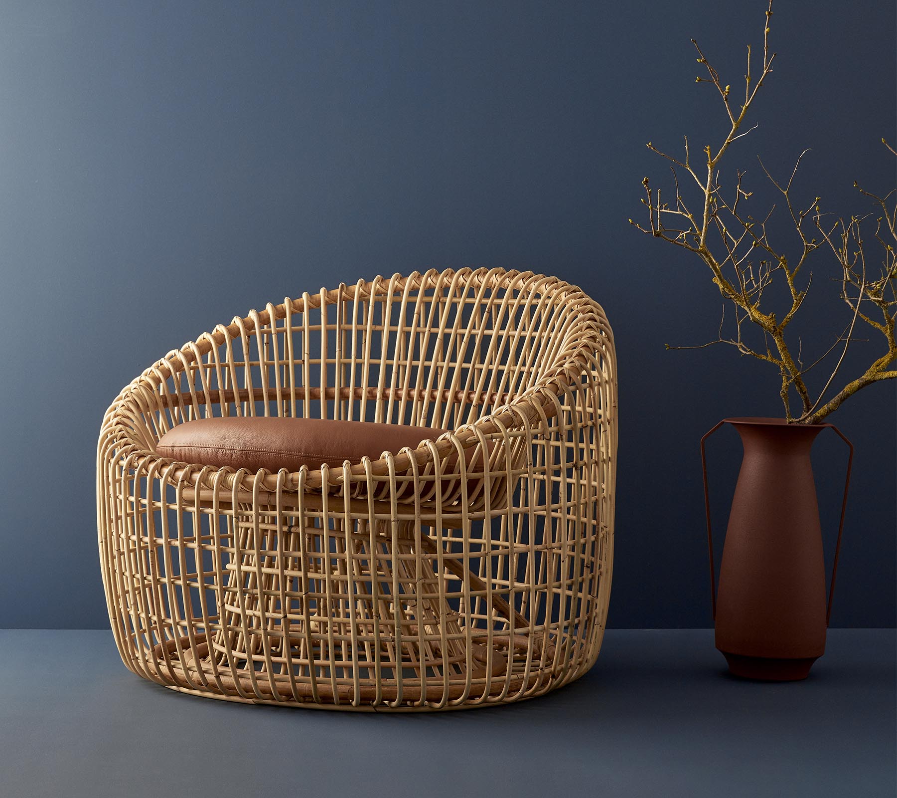 Chaise en osier moderne avec un design arrondi, dotée d'un siège rembourré et complétée par un vase simple avec une brindille.