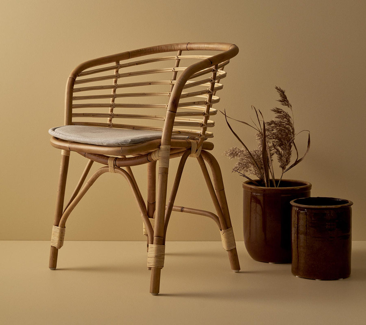 Une chaise en rotin élégante avec un siège rembourré, accompagnée de deux jardinières décoratives avec des plantes séchées.
