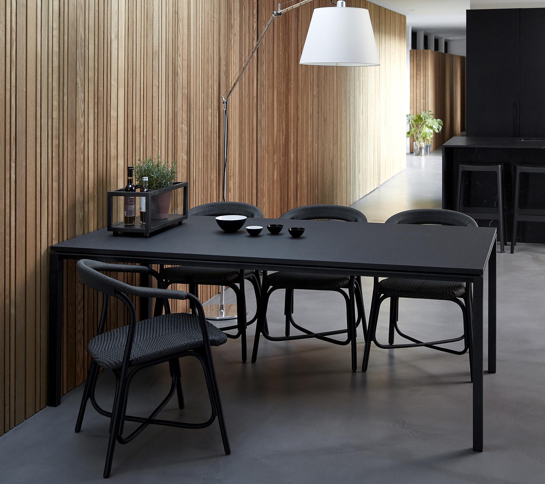 Une table à manger noire et élégante accompagnée de chaises, mise en place avec des bols et des éléments décoratifs, contre un fond de panneaux en bois.