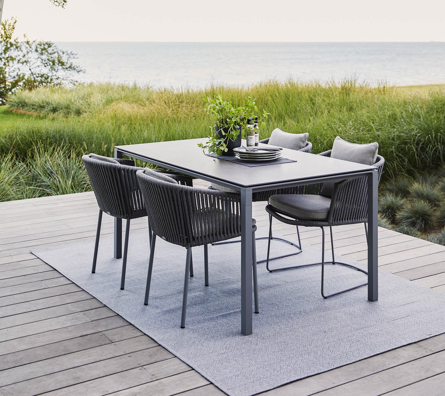Un ensemble de repas en plein air moderne comprenant une table rectangulaire et quatre chaises élégantes, entouré de verdure et d'une vue sereine sur l'eau.