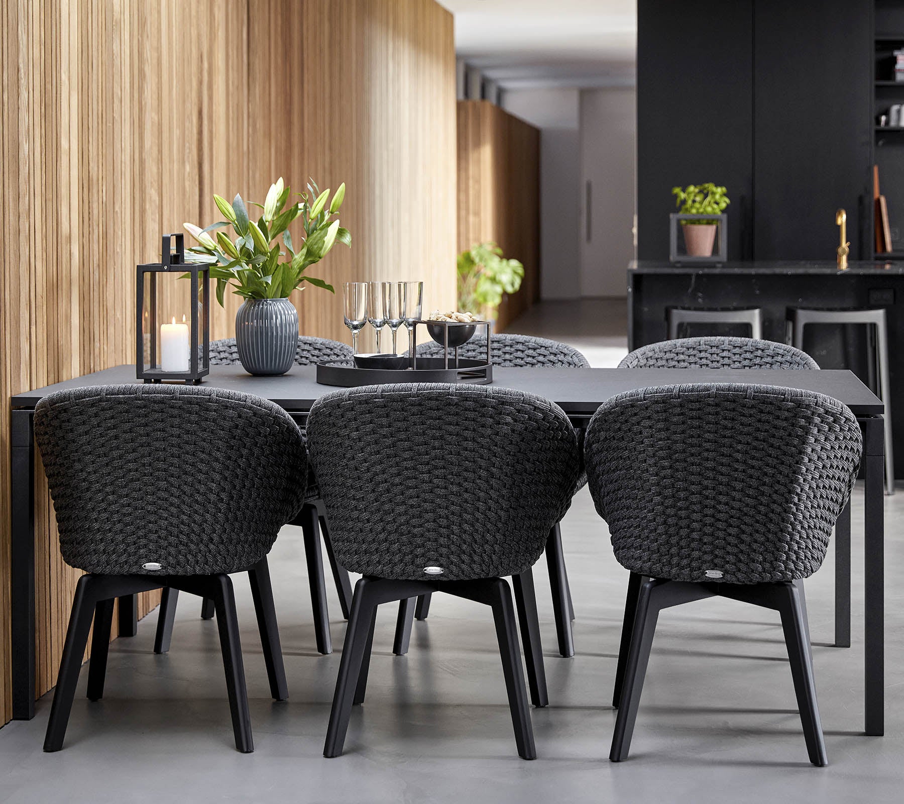 Un aménagement de salle à manger moderne avec une table noire élégante entourée de chaises tissées élégantes, complété par des plantes décoratives et des bougies.