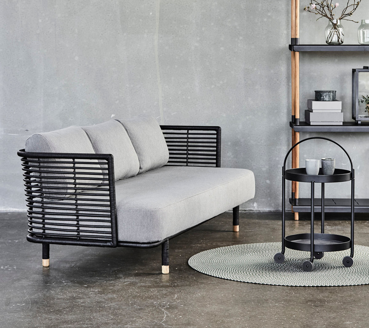 Canapé gris élégant avec des accents en métal noir et des coussins, associé à une table d'appoint ronde sur un tapis texturé contre un fond neutre.