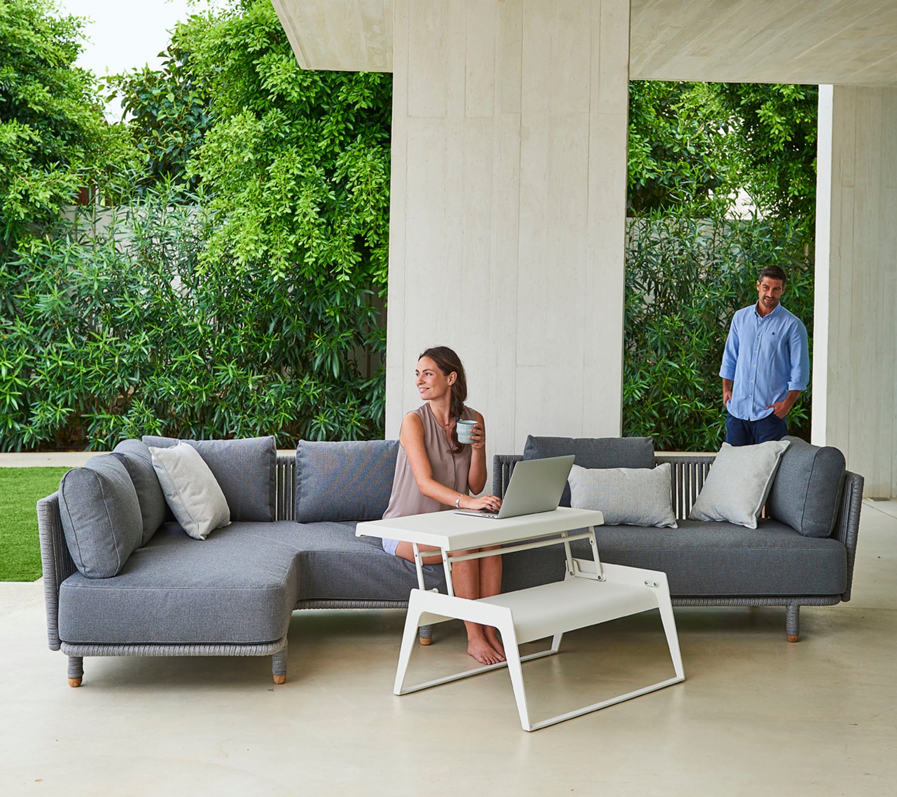 Un canapé sectionnel gris élégant avec des coussins, accompagné d'une table basse moderne, et une femme travaillant sur un ordinateur portable, tandis qu'un homme se tient à proximité.