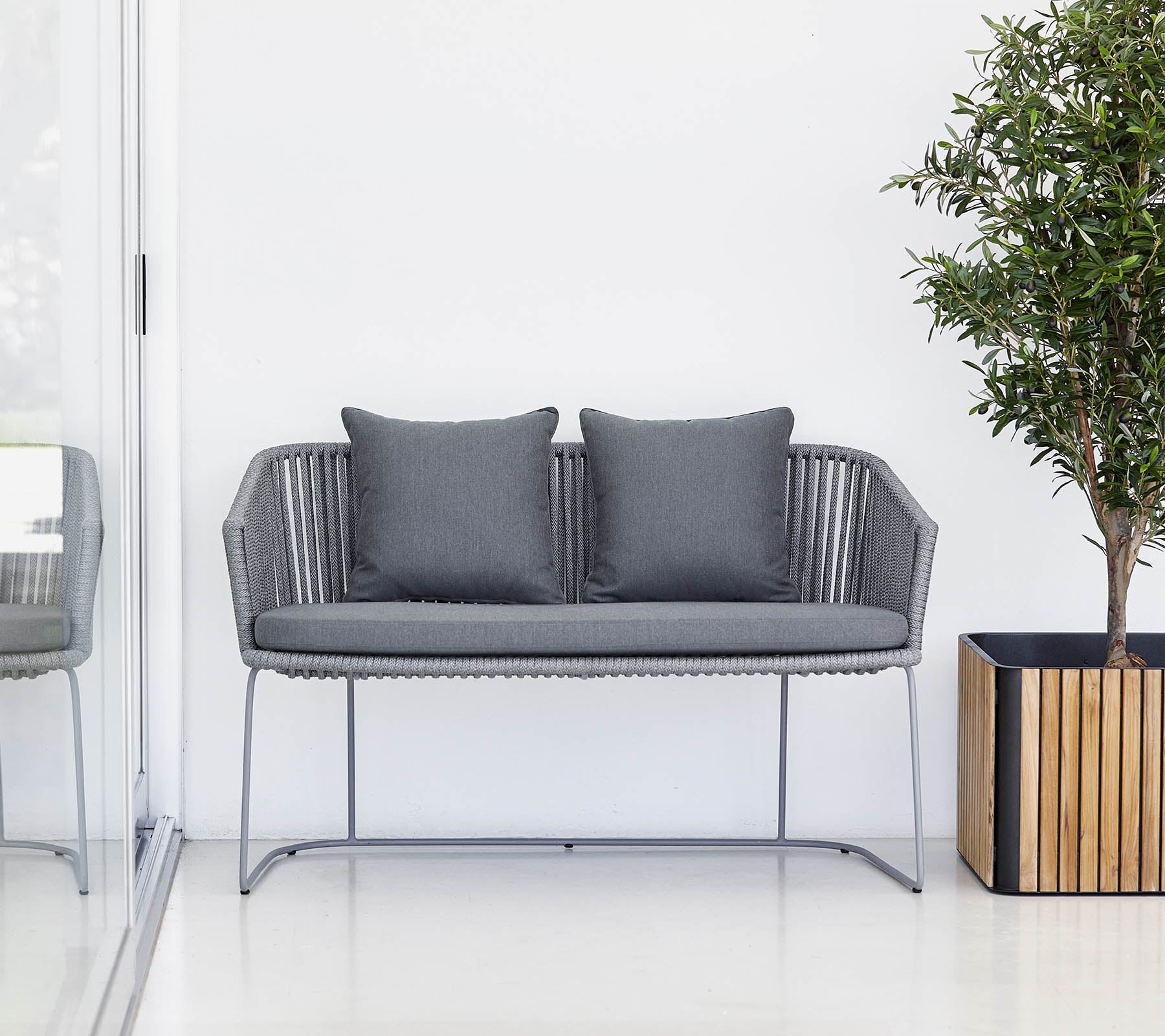 Un canapé moderne gris deux places avec des coussins, accompagné d'une plante en pot et d'un cache-pot en bois.