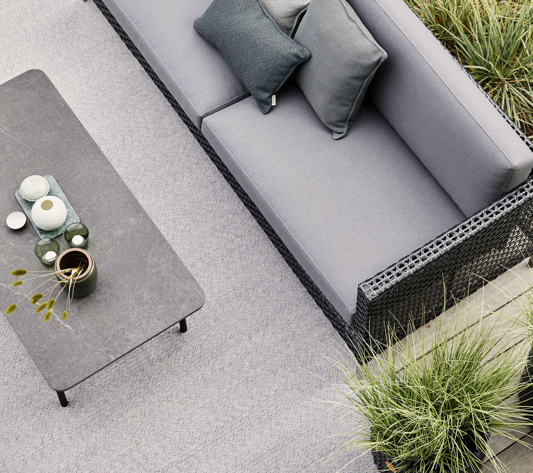 Un canapé gris avec deux coussins se trouve près d'une table minimaliste ornée de céramique et d'une petite plante, entourée de verdure.