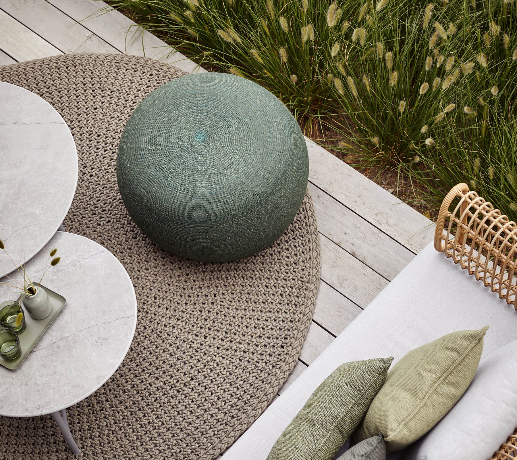 Un espace extérieur confortable avec un pouf rond vert, des tables circulaires et des coussins texturés sur un tapis neutre. Une verdure luxuriante en arrière-plan.