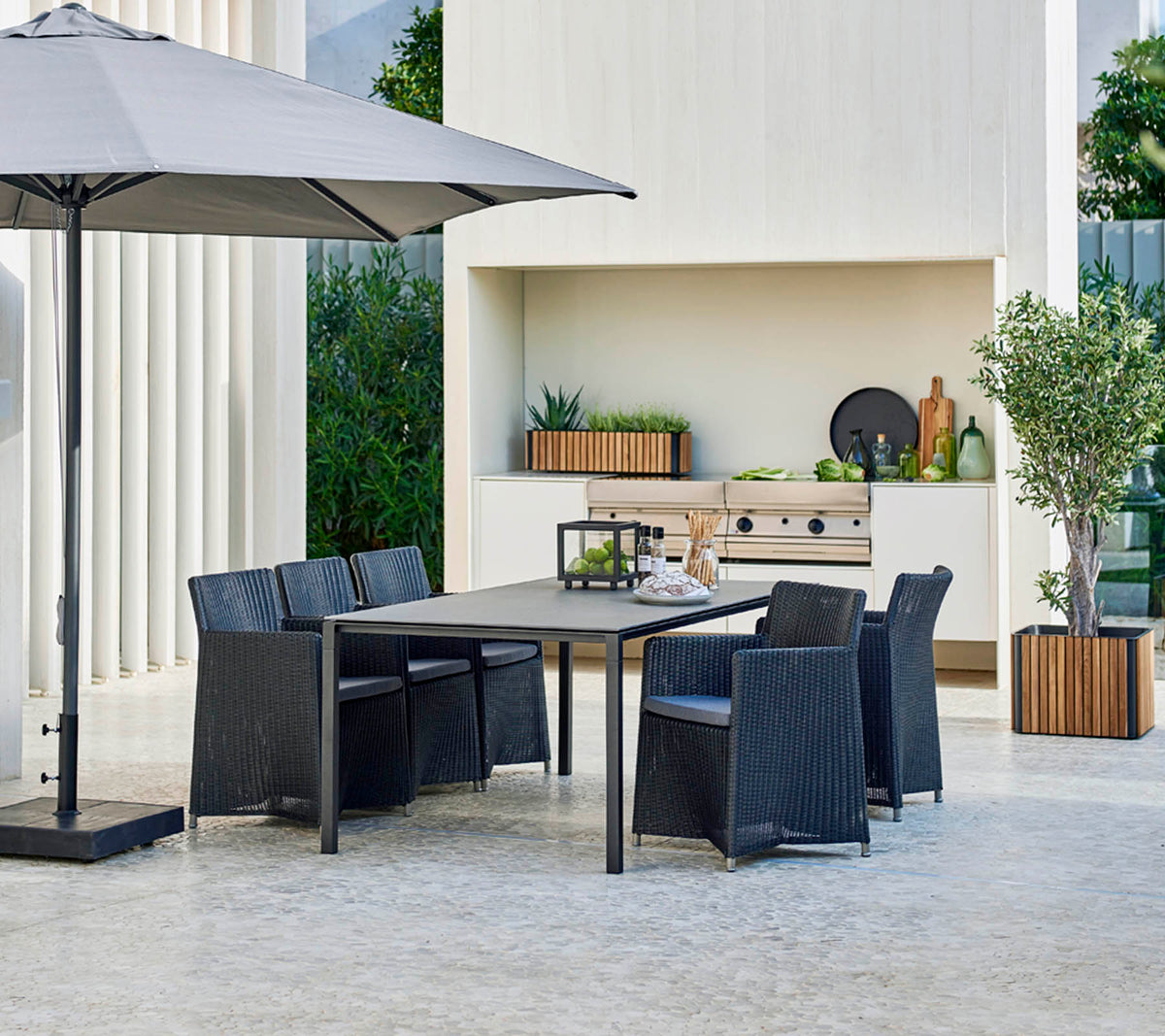 Ensemble de salle à manger d'extérieur moderne avec une grande table, six chaises, un parasol élégant et un arrière-plan avec de la verdure et des éléments décoratifs.