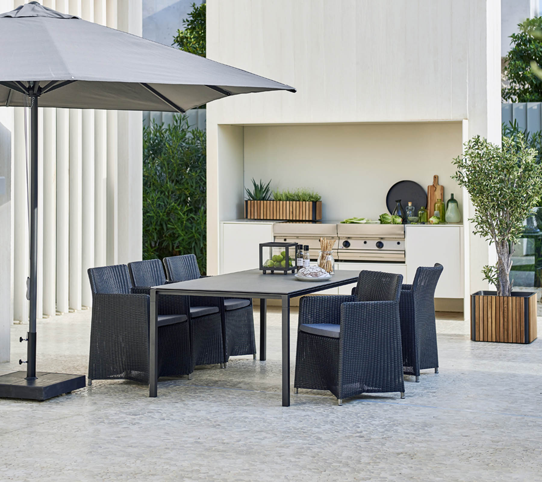 Ensemble de salle à manger d'extérieur moderne avec une grande table, six chaises, un parasol élégant et un arrière-plan avec de la verdure et des éléments décoratifs.