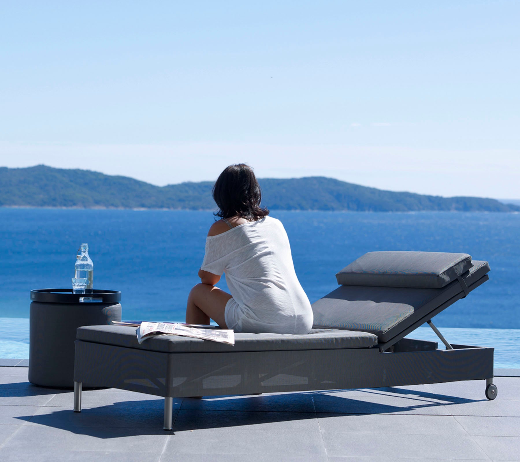 Une femme assise sur un transat, regardant une vue paisible sur l'océan, avec une bouteille d'eau sur une petite table à côté d'elle.