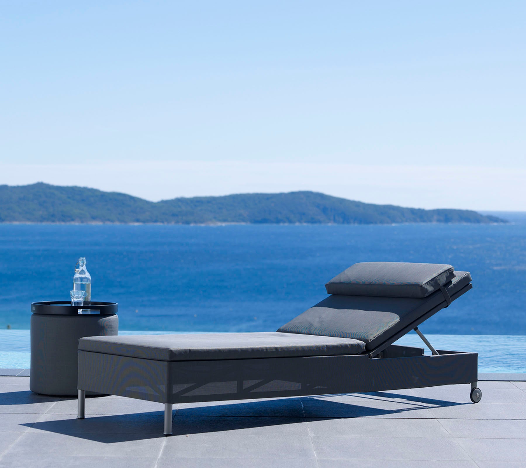 Un fauteuil lounge moderne et une table d'appoint à côté d'un plan d'eau serein avec des collines lointaines sous un ciel dégagé, parfait pour se détendre.