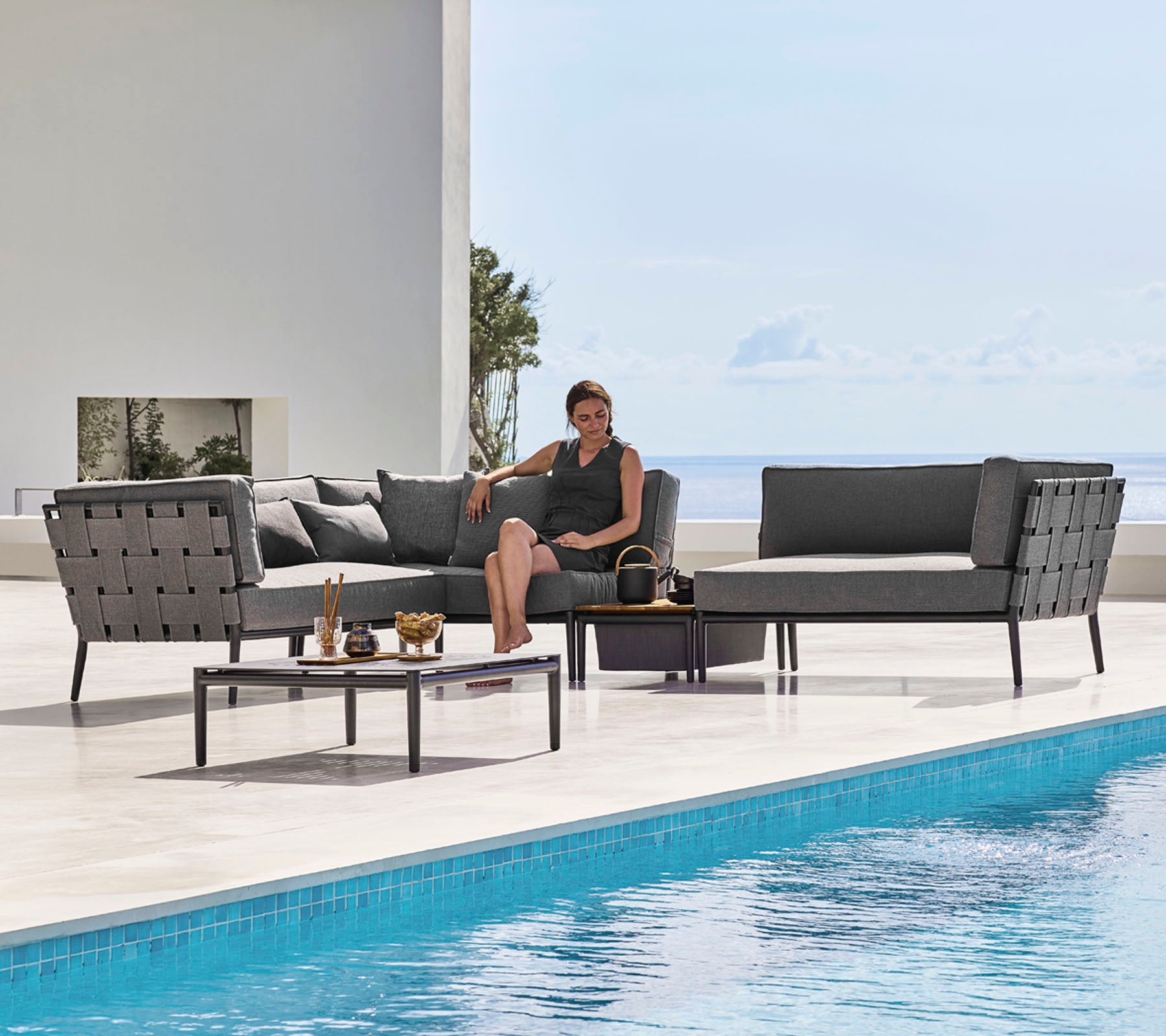 Un ensemble de salon de jardin élégant avec des sièges épurés et une table basse en verre à côté d'une piscine, avec une personne se relaxant confortablement.