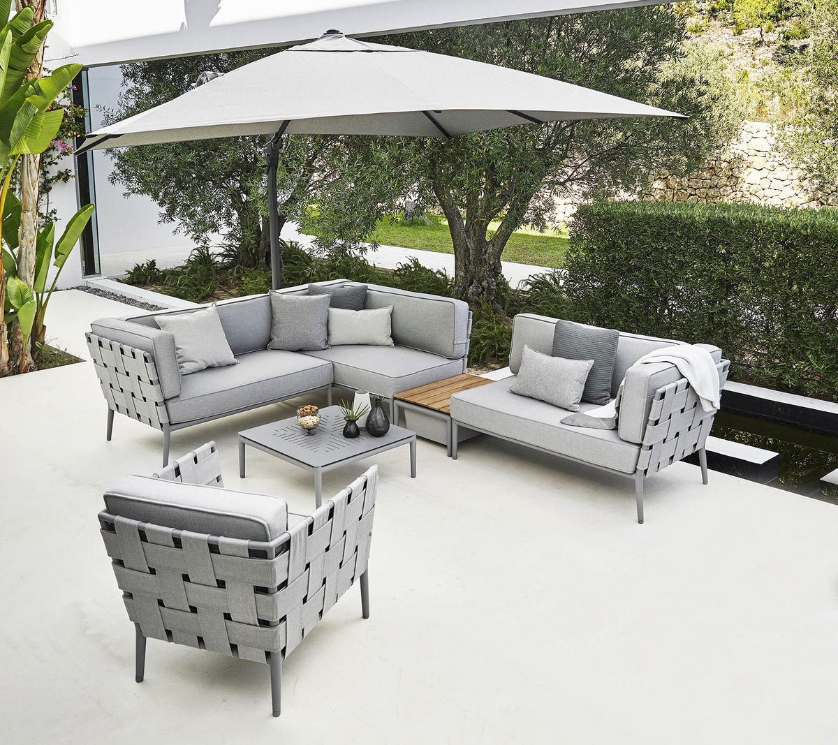 Agencement de sièges d'extérieur élégant avec un canapé d'angle gris, un fauteuil et une table basse sous un grand parasol.