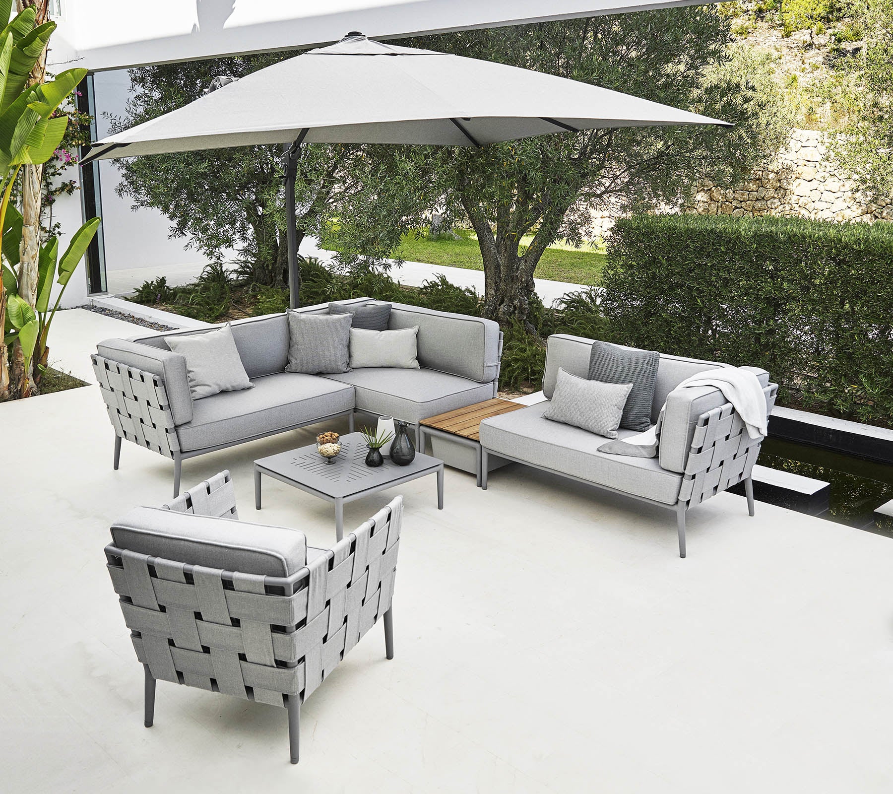 Agencement de sièges d'extérieur élégant avec un canapé d'angle gris, un fauteuil et une table basse sous un grand parasol.