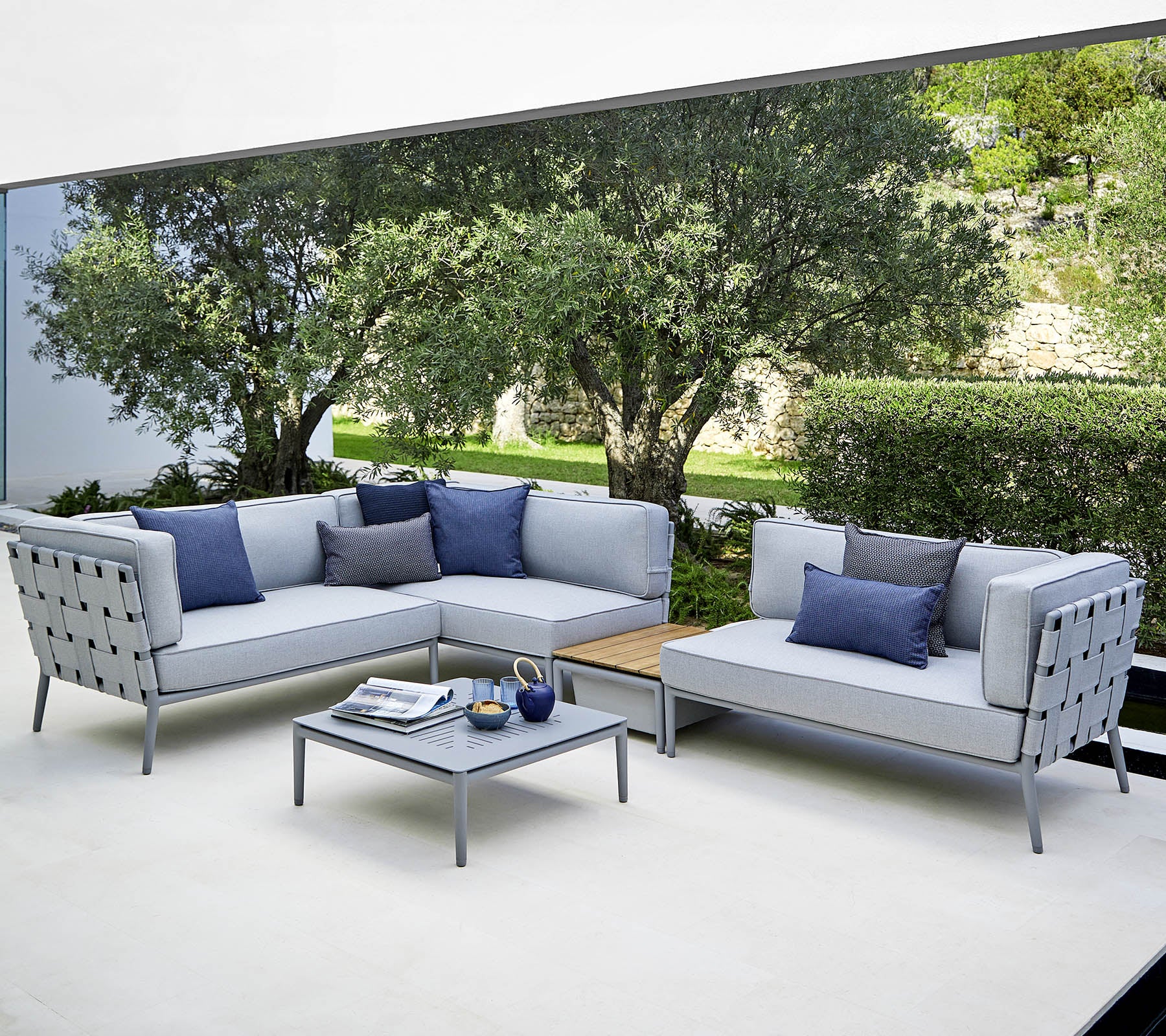 Un aménagement moderne de sièges extérieurs avec des canapés gris clair et des coussins bleus, ainsi qu'une table basse assortie, le tout dans un cadre luxuriant.