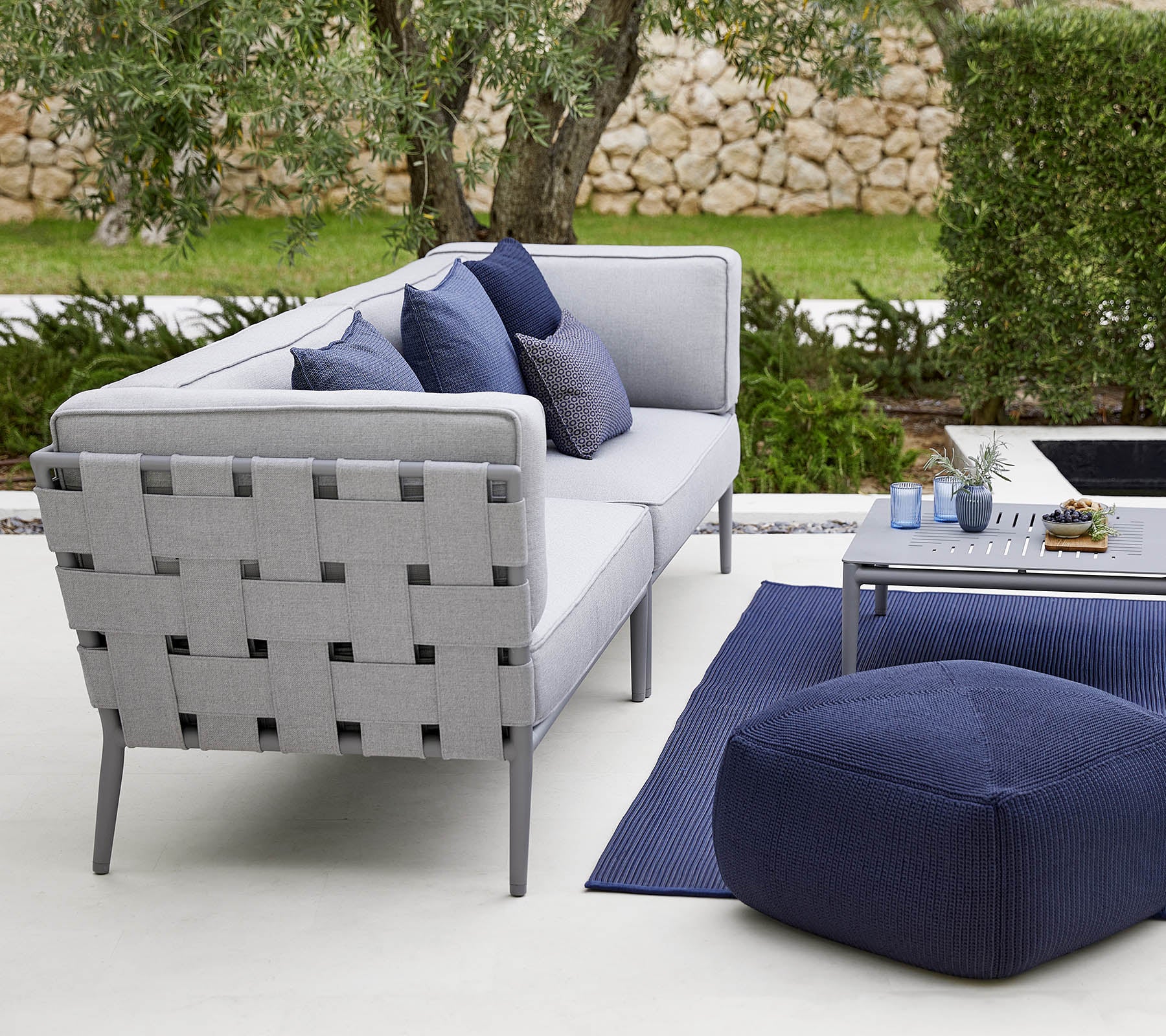 Disposition moderne pour l'extérieur avec un canapé gris accompagné de coussins décoratifs, une table basse élégante et un pouf confortable.